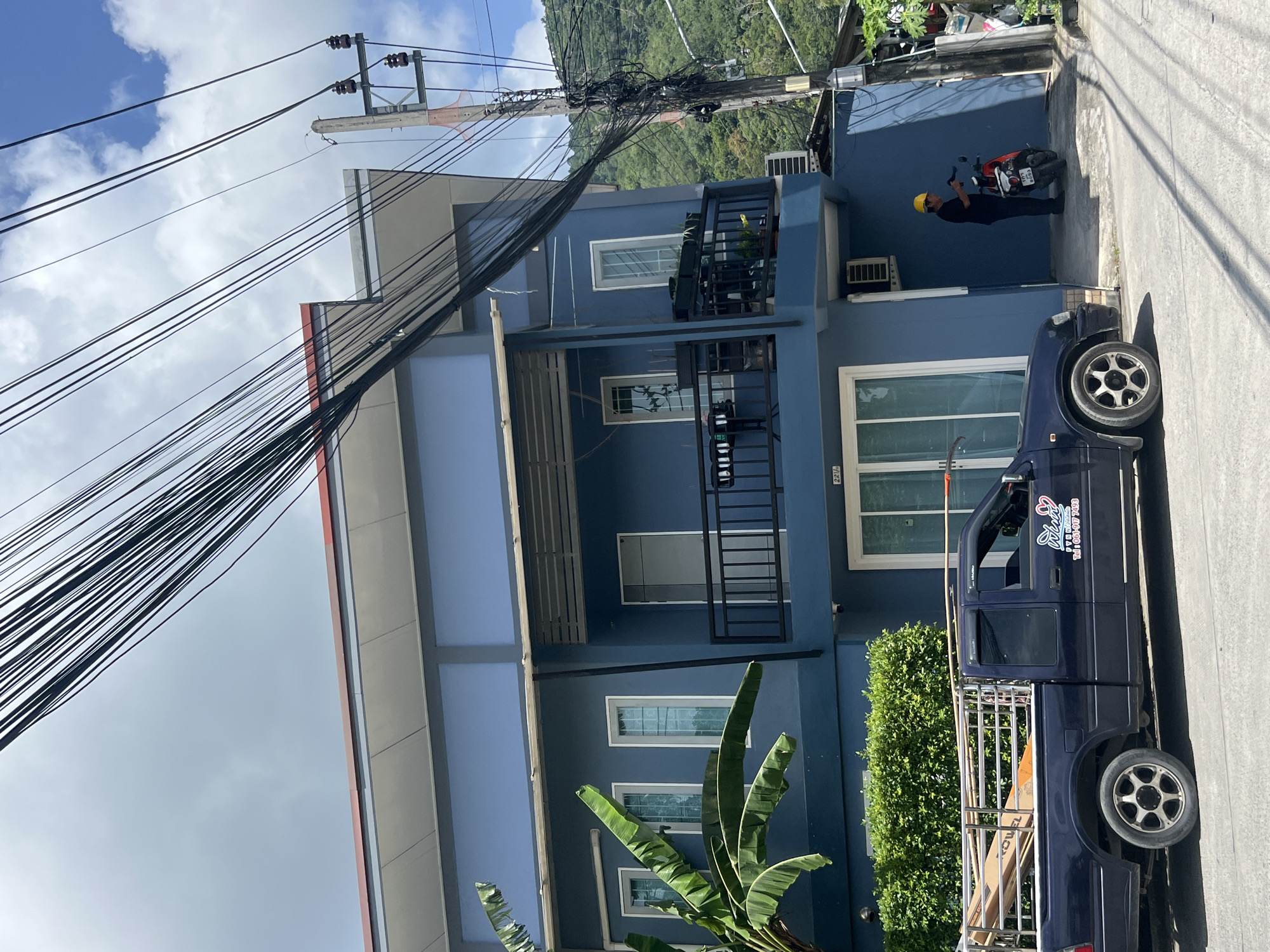 The image depicts a street scene in Choeng Thale, Thailand, with a focus on a blue, multi-story building. A dark blue pickup truck with a silver rack loaded with long, light-colored items is parked on the street in front of the building. The truck has a decal on its side with text and a logo. A person wearing a yellow hard hat and a dark shirt is standing near a red motorcycle in an open doorway of the building. The building has several windows, balconies with black railings, and an air conditioning unit visible on the exterior. Overhead, a dense network of electrical wires and a utility pole dominate the sky. Lush green foliage, including banana leaves from a plant on the left, is visible around the building and in the background, where a hilly, vegetated landscape can be seen. The sky is partly cloudy.
