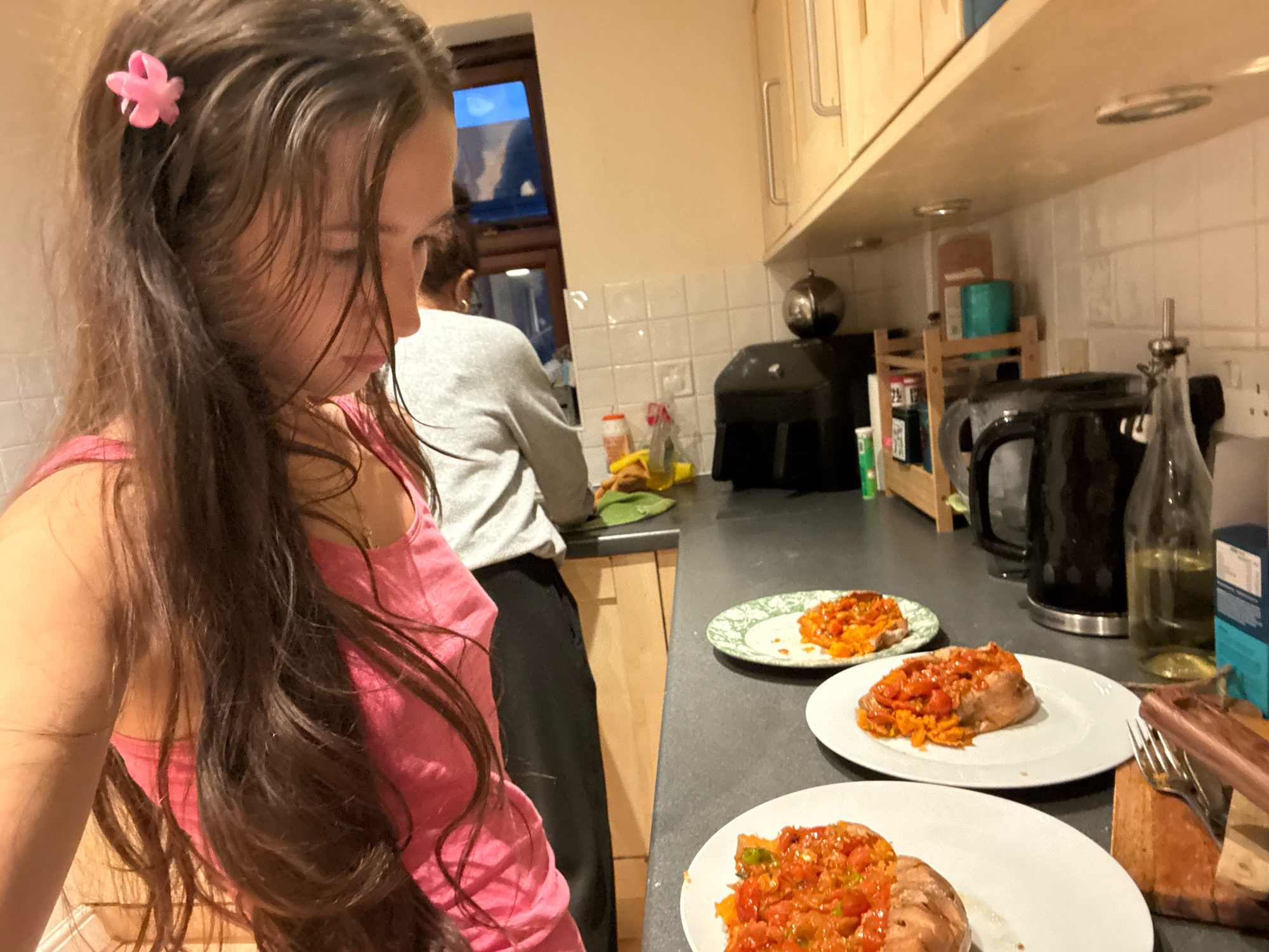 The image depicts a domestic kitchen scene, likely in Reigate and Banstead, United Kingdom, with two individuals present. In the foreground, a young woman with long, dark hair, adorned with a pink flower clip, is partially visible, wearing a pink tank top. Her gaze is directed downwards. In the background, another person, wearing a grey sweatshirt and black trousers, is visible from the back, standing near a kitchen counter. The counter is equipped with a black kettle, a bottle of olive oil, and several plates of food, which appear to be baked sweet potatoes topped with a tomato-based mixture. The kitchen environment features tiled backsplash, wooden cabinets, and a shelf displaying various items. The overall lighting suggests an indoor evening setting.