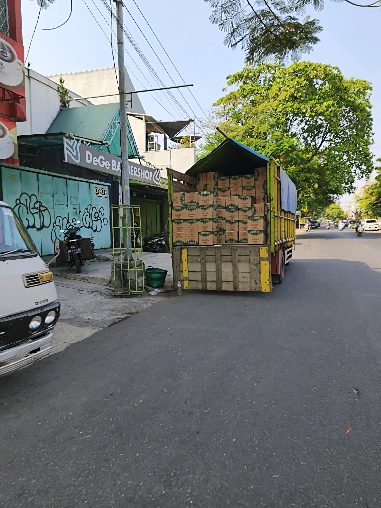 In the city of Surakarta, Indonesia, a yellow delivery truck is parked on the side of a street, its bed filled with stacked cardboard boxes labeled "JUMBO." In front of the truck, a white van is partially visible. Adjacent to the truck, a storefront with a green facade displays graffiti and the sign "DeGe BARBERSHOP." A motorcycle is parked in front of the barbershop. The street is paved, and there are trees and utility poles in the background. The sky is clear and blue.
