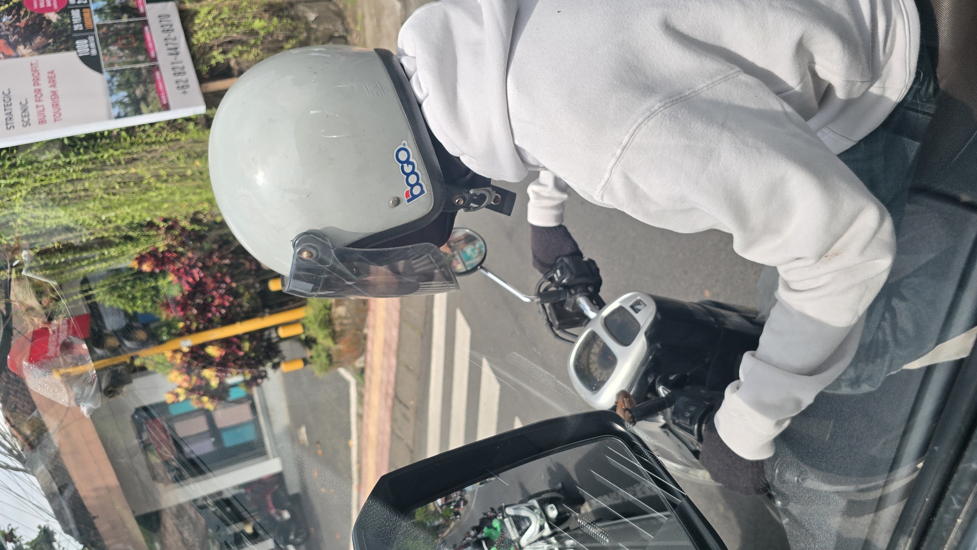 An overhead view, taken through a slightly reflective windshield, captures a person on a scooter or motorcycle in what appears to be Ubud District, Indonesia, during the daytime. The main subject, positioned on the right side of the frame, wears a white hooded sweatshirt and dark gloves. They are donning a light grey open-face helmet adorned with a blue "Booco" sticker and a clear visor. Portions of the scooter's handlebars, dashboard with a speedometer, and a rearview mirror are visible.
The scene depicts a road environment, likely at a stop, with a white crosswalk discernible on the asphalt. A yellow and brown striped curb runs along the roadside. The background features lush green foliage and bursts of red and purple-leafed bushes, suggesting a tropical or semi-tropical setting. A vibrant, multi-colored building (blue, pink, white, brown segments) is faintly visible or reflected in the distance. Reflections on the windshield include what looks like a red plastic object and outlines of other vehicles.
On the far left, a tall white sign with black text is partially visible. Readable phrases and numbers on the sign include "+62 821-4472-8370" (an Indonesian phone number), "STRATEGIC.", "SCENIC.", "BUILT FOR PROFIT.", "TOURISM AREA.", "1000", and "25 YEARS LIFESPAN". The bright lighting indicates sunny weather.