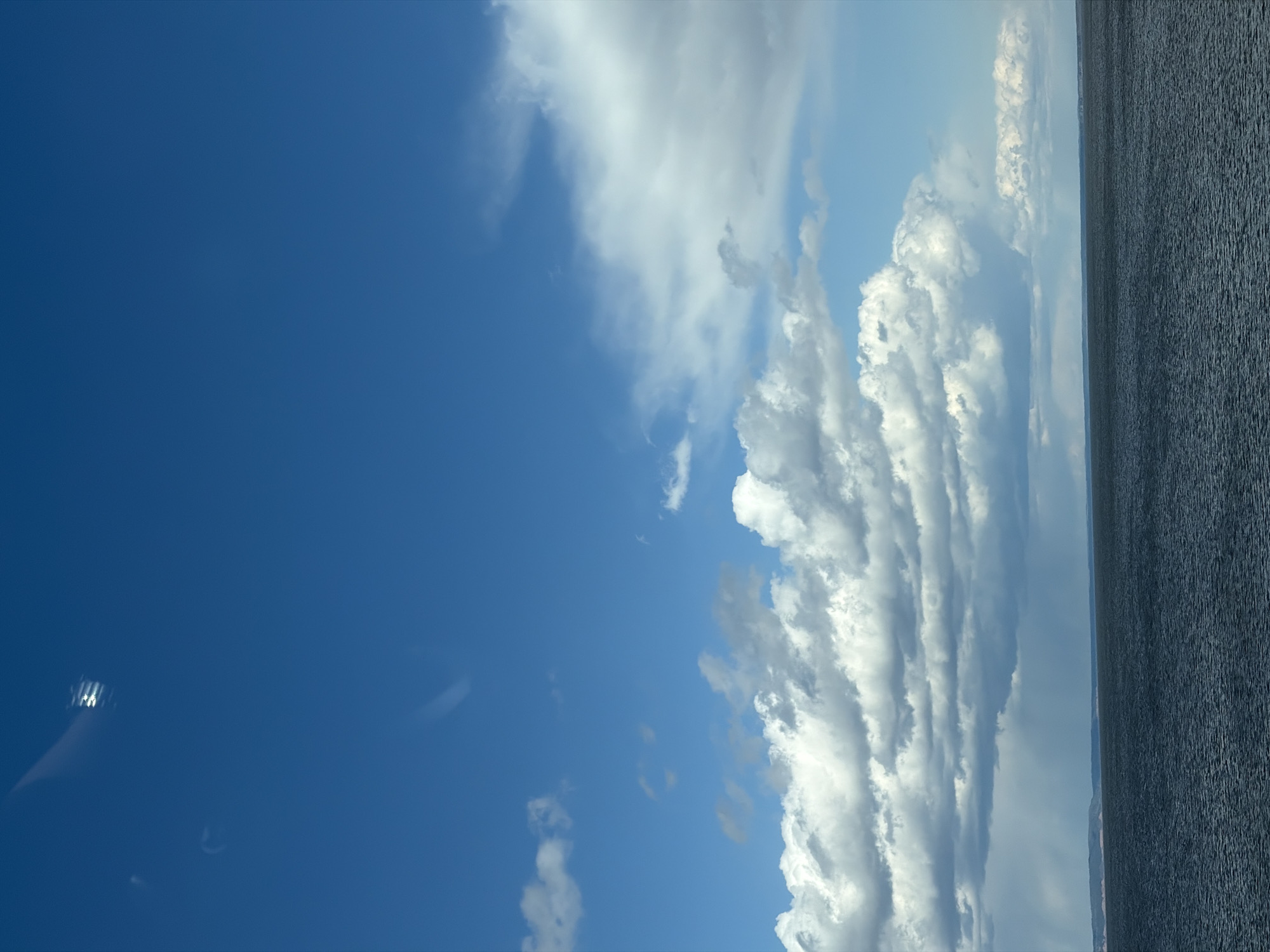 This image presents a vast outdoor landscape featuring a dramatic sky above a large body of water. When viewed in its correct orientation, the horizon line clearly separates the expansive sky, which occupies roughly the upper two-thirds of the frame, from the water in the lower third.
The sky is a rich, deep blue, filled with an array of cloud formations. Prominent among these are large, brightly lit white cumulus clouds, some arranged in an impressive, almost layered pattern that stretches across the middle of the sky. Interspersed with these are more diffuse, greyish clouds, indicating varying atmospheric conditions. The intense brightness of the white clouds suggests a sunny daytime setting.
The lower portion of the image reveals a large body of water, appearing a deep, dark greyish-blue. Its surface is textured with numerous small ripples, indicating a light breeze. A faint, low-lying landmass or distant shoreline can be discerned along the horizon line, particularly towards the right.
The scene depicts a calm and expansive natural environment, likely captured during the day with generally clear weather. Given the context of Chaikovsky, Russia, the body of water is likely a segment of the Votkinsk Reservoir. There are no visible people, human-made objects, specific activities, or text present in the image.
