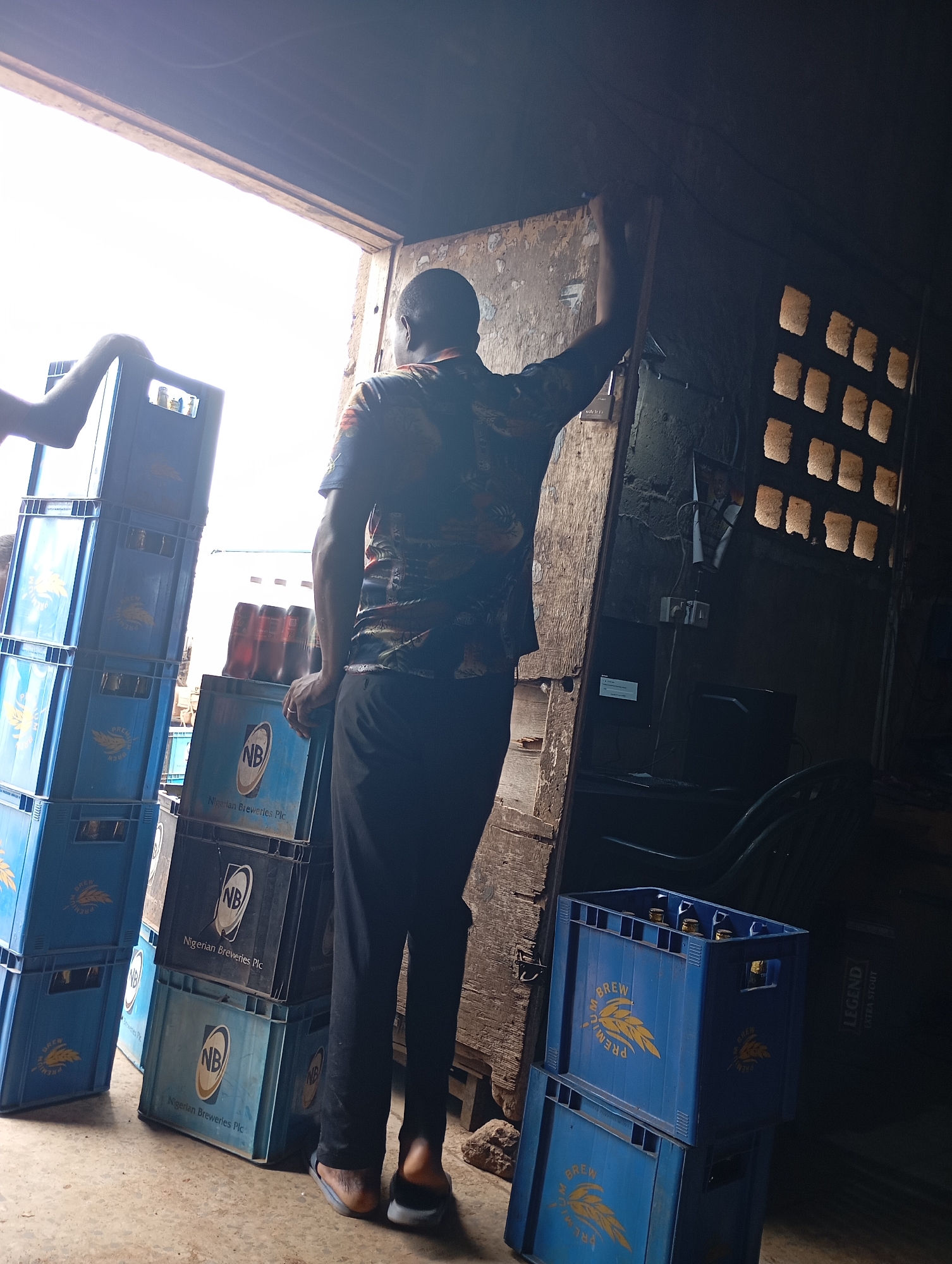 In Shendam, Nigeria, a man stands inside a dimly lit structure, possibly a warehouse or workshop, with a large, open doorway revealing a bright exterior. He is looking out of the doorway, with his back mostly to the camera. He is wearing a colorful, patterned shirt and dark trousers. To his left, stacks of blue plastic crates, some labeled "BREW PREMIUM" and "Nigerian Breweries Plc," are piled high. These crates appear to contain bottles, likely beer. To the right of the man, the interior wall is textured, with a window featuring several square openings that allow light to filter through. Electrical outlets and wires are visible on the wall, and a dark piece of furniture, perhaps a chair, is partially visible in the background. The floor is concrete.