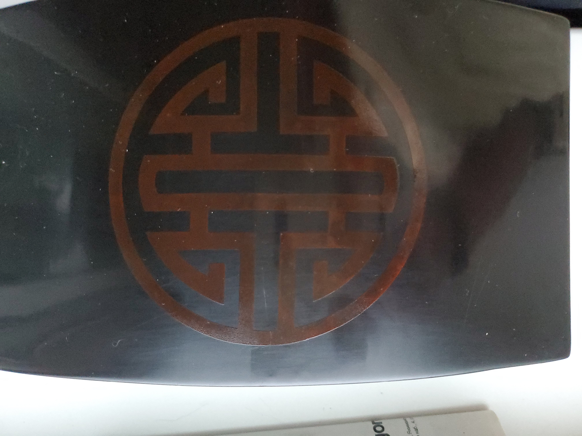 The image shows a close-up of a dark, rectangular object with rounded corners. A circular emblem with intricate, geometric brown patterns is centrally placed on its surface. The emblem appears to be a stylized knot or a symbol with multiple interconnected lines. The object has a glossy finish, and there are some minor scratches and dust specks visible on its surface. To the right of the object, a small portion of a white paper with some text is visible. The lighting suggests an indoor setting. The image does not provide any information about the location in Tiraspol, Moldova.