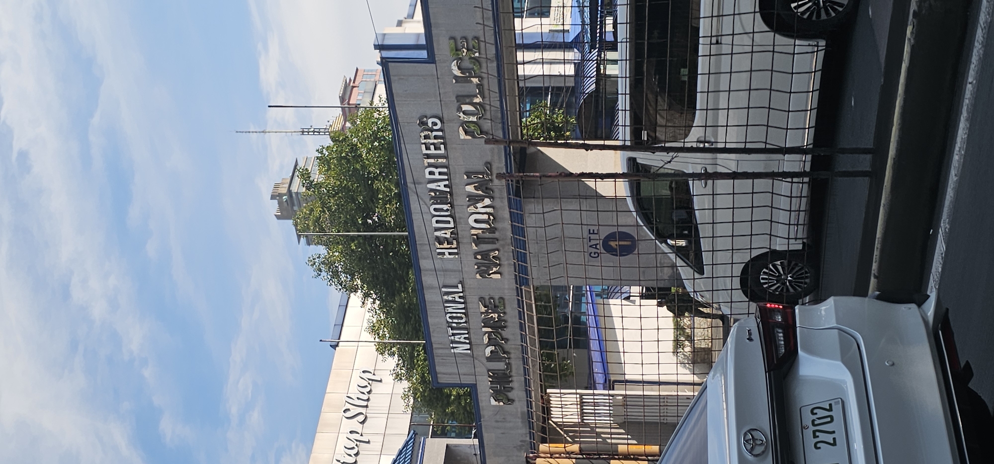The image shows the National Headquarters of the Philippine National Police in Quezon City, Philippines. The signage reads "NATIONAL HEADQUARTERS PHILIPPINE NATIONAL POLICE." A gate, labeled "GATE 1" with a blue circle and the number "1" inside, is visible. A white van is parked inside the gate, and a person in what appears to be a uniform is standing near the gate. A white car is parked in the foreground, facing away from the camera. The background shows buildings and trees under a partly cloudy sky.