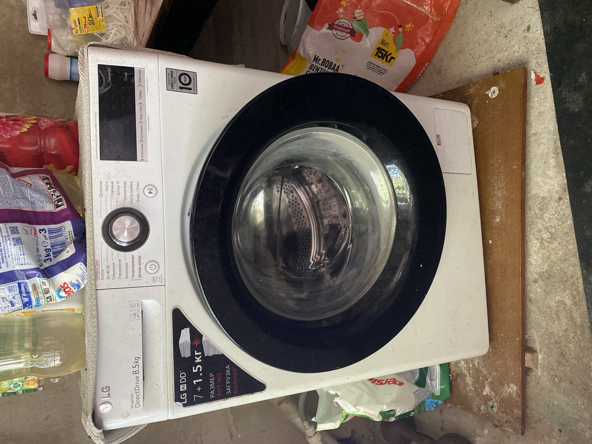 A front-facing, medium shot captures a white LG washing machine situated on a wooden plank, which itself rests on a concrete floor. The machine is positioned indoors, likely in a storage area or utility room, given the presence of various bags of laundry detergent and other household items in the background. The washing machine displays a digital interface and a control panel with several program options. The drum of the washing machine is visible through the glass door. The overall scene suggests a functional, though slightly cluttered, domestic setting. The location is identified as Shakhtinsk, Kazakhstan.