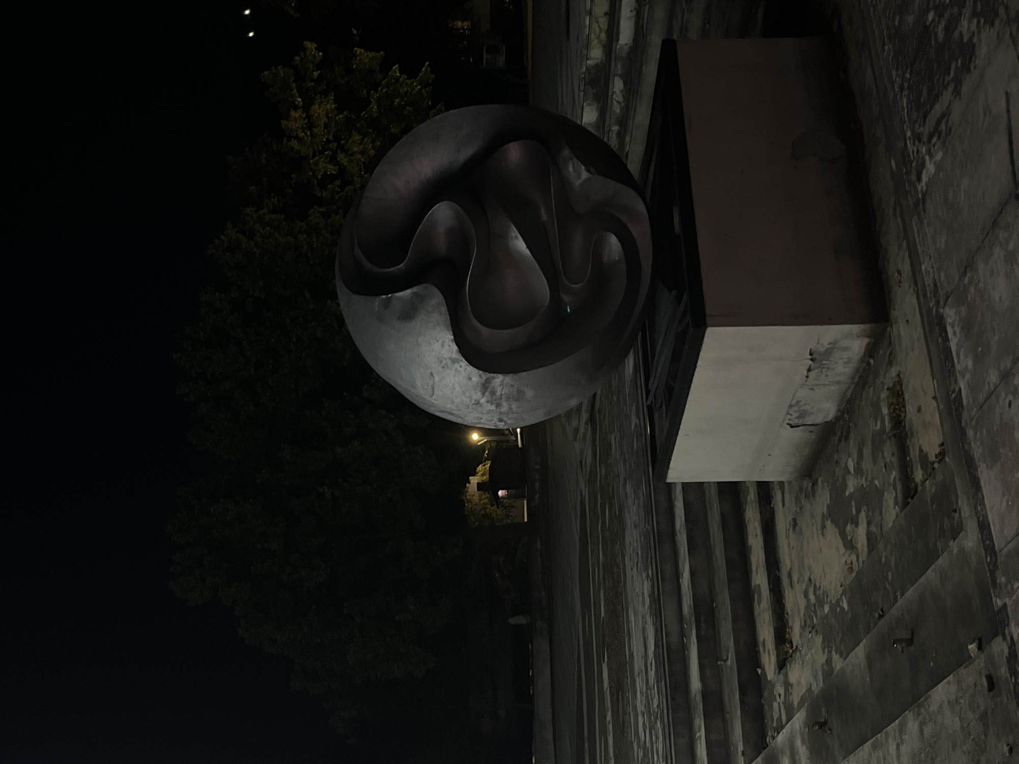 The image shows a large, dark, abstract metal sculpture in an outdoor plaza at night. The sculpture is spherical in shape with swirling, organic forms cut into its surface, creating negative space within. It is mounted on a rectangular, light-colored pedestal, which appears worn and damaged in places.
The plaza itself is made of concrete and features tiered levels with steps. The concrete is cracked and weathered, with some debris and fallen leaves scattered across the surfaces, suggesting neglect or disuse.
In the background, silhouetted trees are visible against the dark night sky. A streetlamp with a warm yellow glow illuminates a small building or structure behind the sculpture. Another fainter light source is visible further to the right. The overall atmosphere is somber and somewhat desolate, illuminated by artificial light in the darkness.
This sculpture is located in Vratsa, Bulgaria, specifically in the area of the National Revival House of Culture "Prosveta" which is adjacent to the Vratsa History Museum and the Municipal Administration building. The plaza is part of the pedestrian zone in the center of the city.