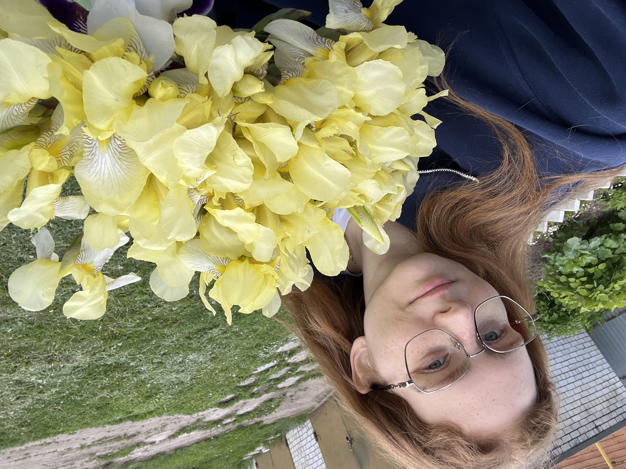 The image is a brightly lit outdoor portrait, likely a selfie, of a young woman holding a large bouquet of yellow irises. The photograph is oriented with the subject's head tilted towards the bottom left, appearing rotated 90 degrees clockwise from a standard portrait.
The main subject is a young woman with long, wavy reddish-brown hair and visible freckles across her face. She is wearing modern, thin-framed silver glasses and has a calm, neutral expression. She is dressed in a dark blue garment, possibly a sweatshirt or jacket, and a delicate silver chain is visible around her neck.
She is holding a dense bouquet of numerous yellow irises. The petals are predominantly a soft, pale yellow. Closer inspection reveals that some of the inner petals, particularly the 'falls' (lower petals), are lighter, almost white, with distinct brown or purple striped patterns.
The background suggests an outdoor setting, likely a garden or yard in Новая Тура, Russia. To the left (when viewed upright), there is lush green grass and a dirt or gravel path. A light-colored brick structure, possibly a wall or building, is partially visible. To the right (when viewed upright), there is green foliage from bushes or trees, and a grey, textured surface that appears to be a roof or another building element. The bright, even lighting indicates it is daytime, likely a clear day. No visible text is present in the image.