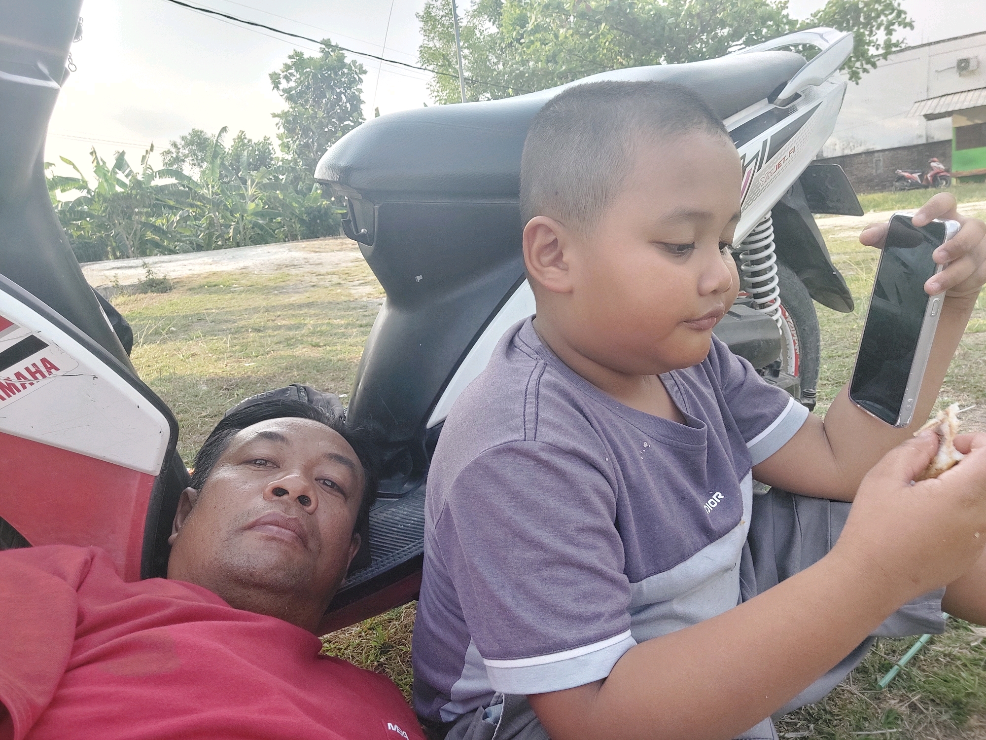 An adult male wearing a red shirt and a young boy wearing a purple and grey t-shirt are in the foreground, lying on the ground next to a white and red motorcycle. The man is on the left, looking directly at the camera. The boy is on the right, holding a smartphone and a piece of food. In the background, there are banana trees, grass, and a building with a red and green roof. The image appears to be taken in an outdoor setting, possibly in Sarangan, Indonesia, given the vegetation and architecture. The overall lighting suggests it is daytime.