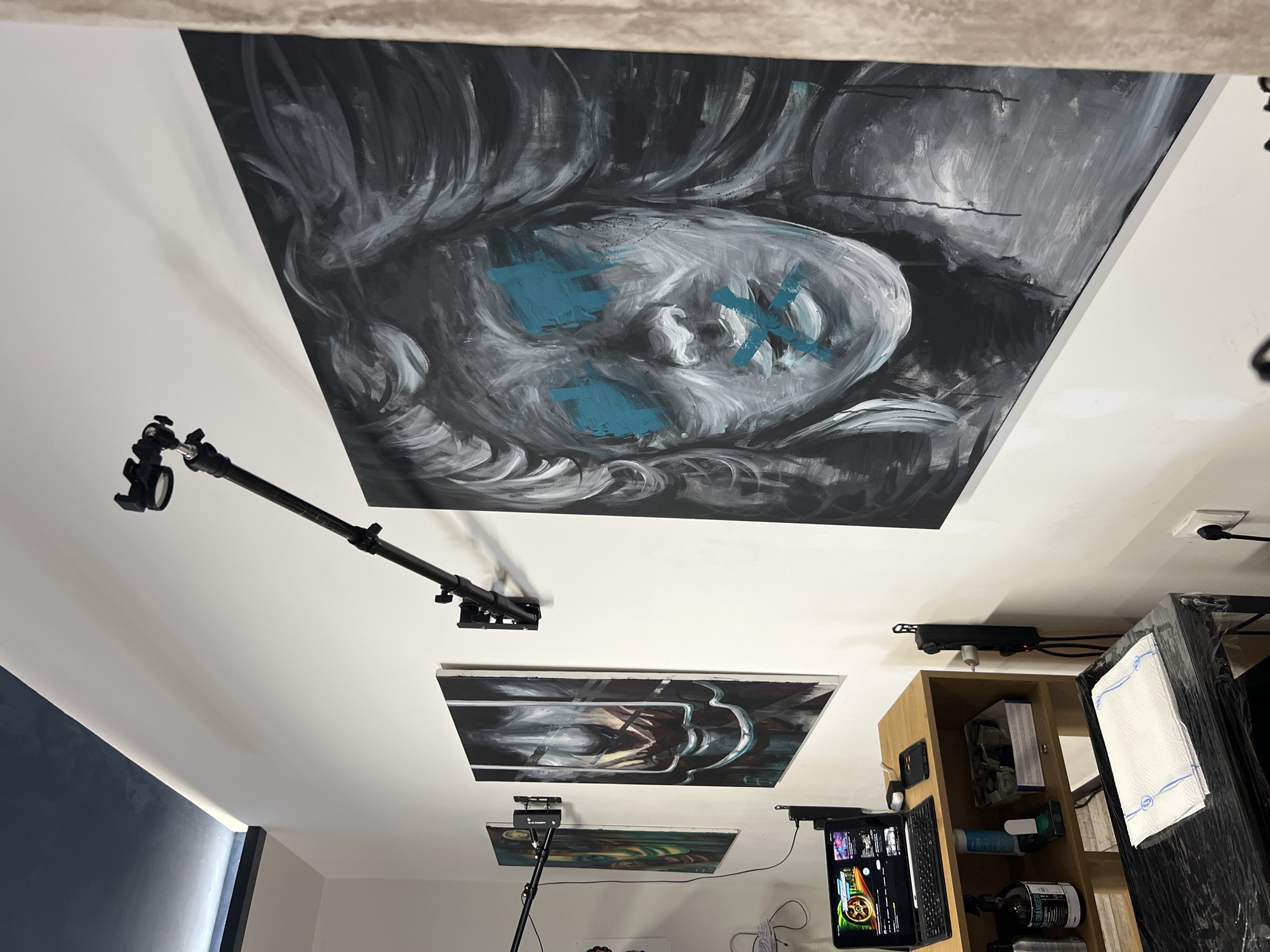 The image shows an interior space with a focus on the ceiling. Three framed artworks are affixed to the ceiling. The largest, topmost artwork depicts a monochrome portrait with blue "X" marks over the eyes and mouth. Below this, another artwork shows a darker, abstract design. The third and smallest artwork is visible further down the scene. A black boom stand extends from the side wall toward the upper artwork, ending with a camera mount. The side wall is painted a dark blue color. A wooden shelving unit sits against the other wall holding an iPad, keyboard, and various items, including a black power strip mounted above it. A wrapped object stands to the right of the wooden shelf, and the overall environment is brightly lit. The location is not discernible from the provided image without additional context clues.