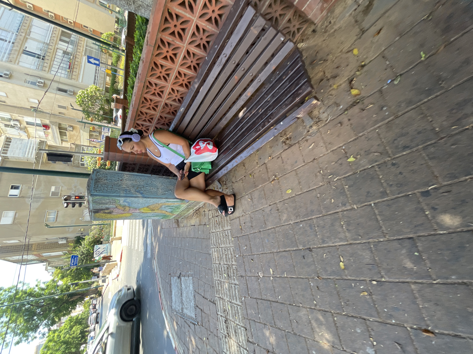 A woman with light-colored hair and headphones sits on a wooden park bench on a sidewalk. She is wearing a white tank top, black shorts, and black sandals. A green bag is on her lap, and a white and red bag is beside her on the bench. The bench is positioned in front of a decorative brick wall. The sidewalk is made of grey pavers. In the background, a street is visible with cars, traffic lights, and buildings. The image appears to be taken on a sunny day. The location is Netanya, Israel, specifically near the intersection of Rehovot Street and Yehuda Halevi Street.
