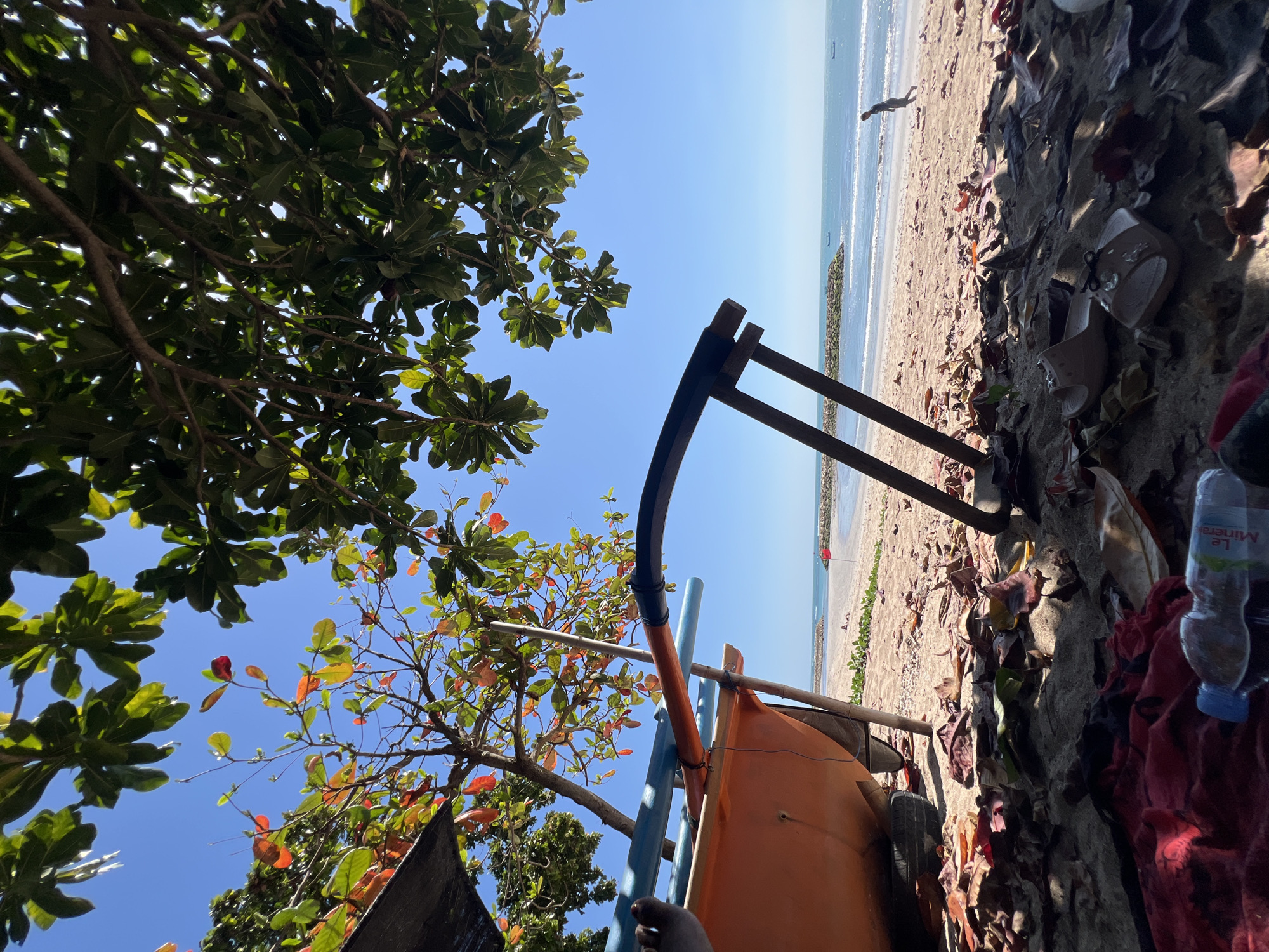 A low-angle shot captures a beach scene in Kuta, Indonesia, on a clear, sunny day. In the foreground, the hull of an orange boat rests on the sand, propped up by wooden poles. To the left, a person's bare foot is visible, suggesting they are sitting near the boat. The sand is littered with fallen leaves. Further back, a calm ocean stretches to the horizon, with a few boats visible in the distance. A person can be seen jogging along the shoreline, and a red flag is planted on the beach. Lush green trees with some autumn-colored leaves overhang the scene from above.