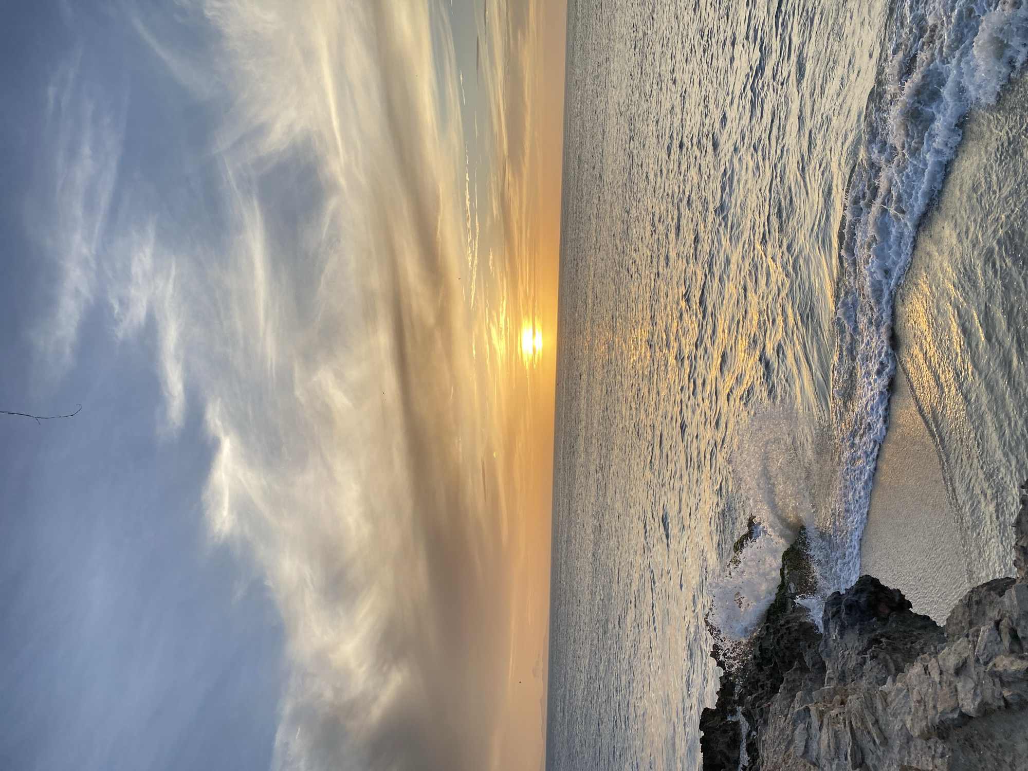 The image depicts a sunset over the ocean in Juan Griego, Venezuela. The sky is a mix of blue and golden hues, with wispy clouds streaked across it. The sun is positioned on the horizon, casting a bright glow over the water. The ocean surface is textured with small waves, reflecting the sunlight. The image is taken from an elevated position, showcasing rocky outcroppings in the foreground. Waves are crashing against the rocks. There is no evidence of people.