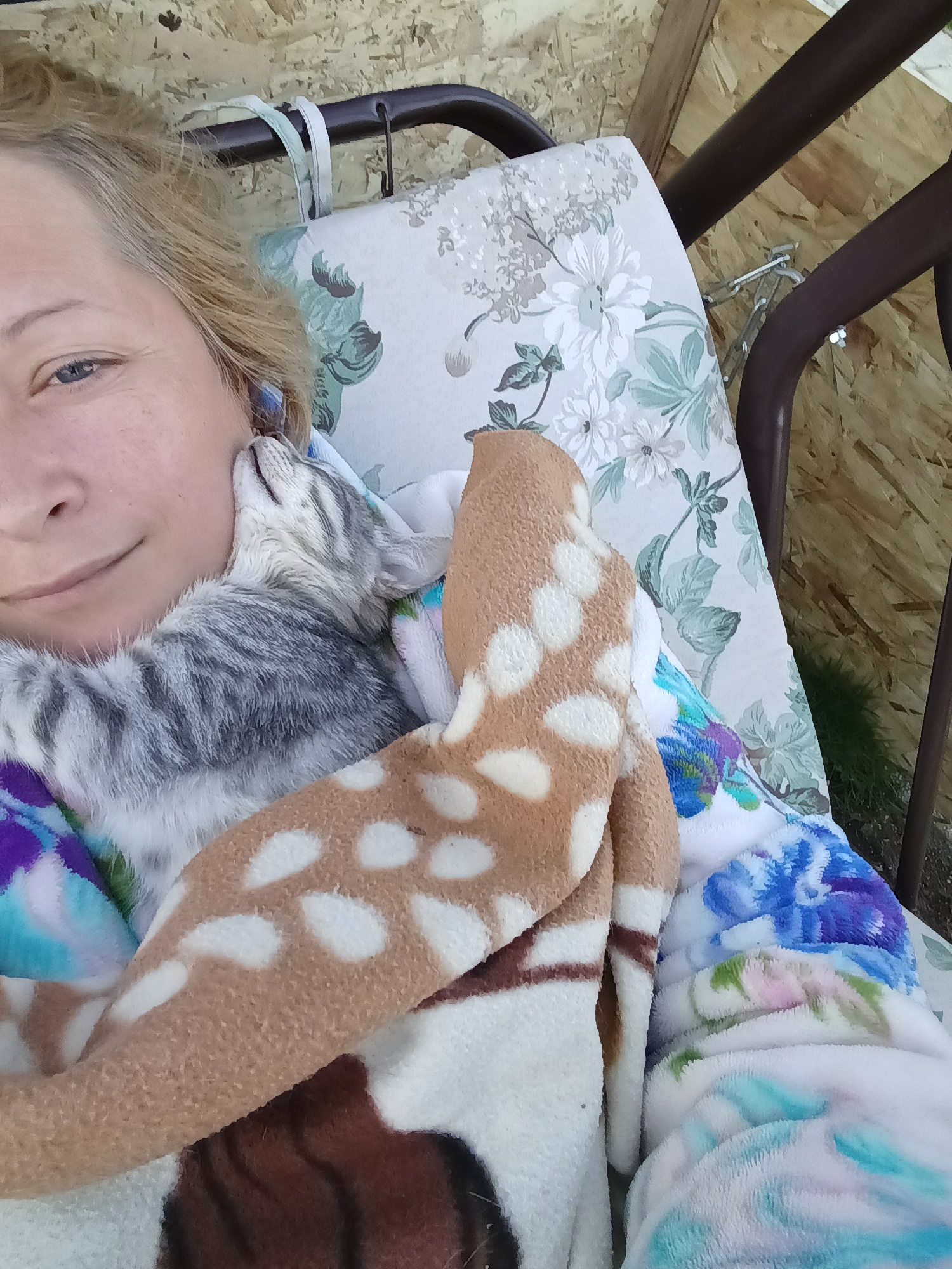 A blonde-haired person lies on a floral-patterned swing seat, holding a grey tabby kitten. The person's face is visible, looking towards the camera with a slight smile, with the kitten nestled against their cheek. A blanket with a fawn pattern is draped over the person and kitten, along with another blanket with a blue and white floral design. The background consists of particleboard walls and a dark metal swing frame. The scene appears to be outdoors, possibly on a porch or in a garden area, in Ivanovskoe, Russia.