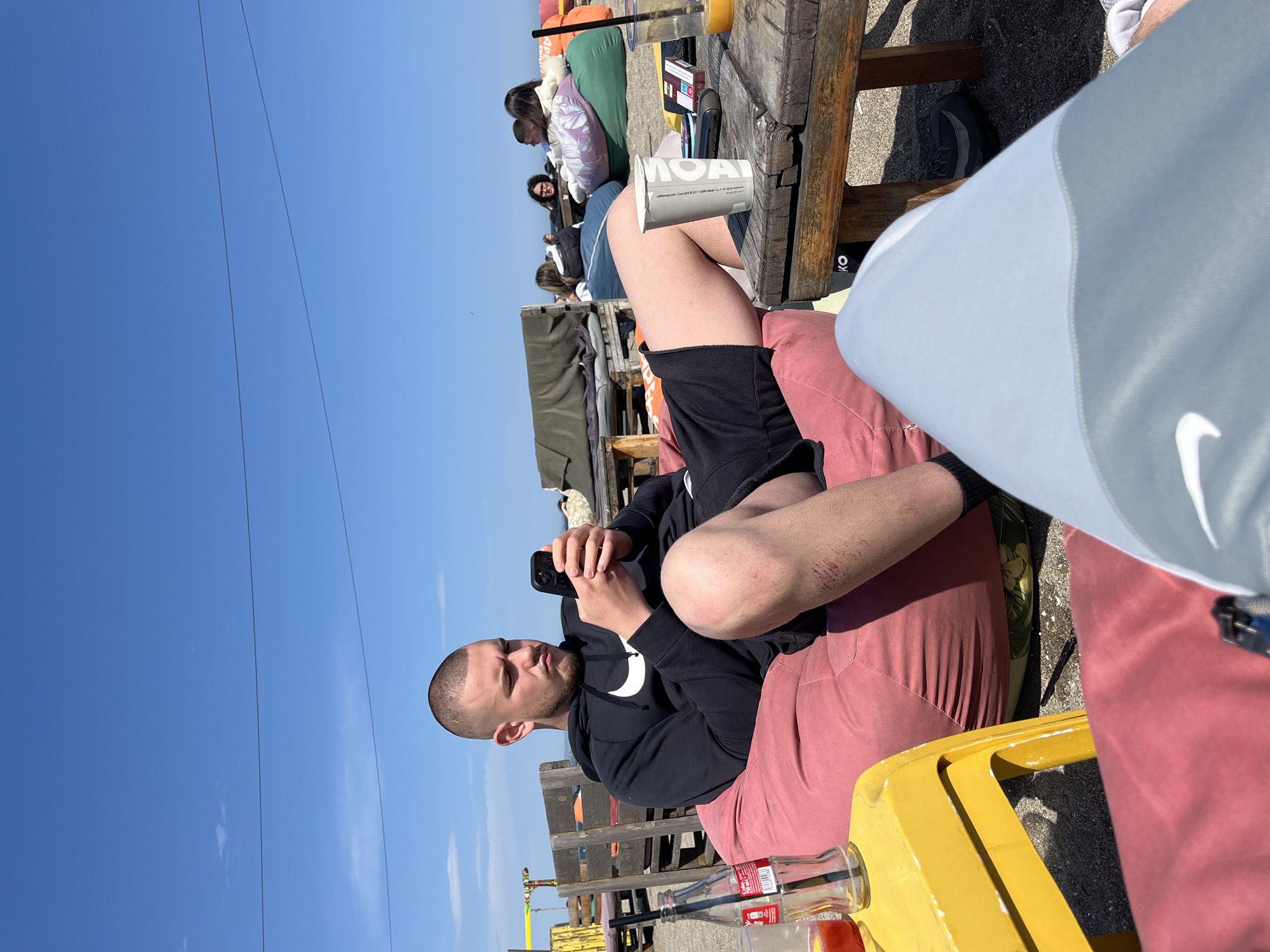 The image captures an outdoor, sunny scene, likely at a casual cafe or lounge area. The primary subject is a man in the foreground, seated on a reddish-pink beanbag or cushioned seat. He has short hair and a beard, and is wearing a black hoodie with a white Nike swoosh logo on the front, and black shorts. He is looking down at and interacting with his black smartphone, held in both hands. Visible scratches or scabs are present on his right knee/shin area. His legs are casually crossed or bent, resting on the seat.
To the man's right, a rustic wooden table holds a white disposable cup with the text "MOAI" printed on it, and a glass containing an orange-colored drink. Near his left leg, a clear glass Coca-Cola bottle, partially filled with a reddish drink, rests on a yellow surface, possibly another low table or tray.
In the mid-ground and background, other patrons are visible, sitting at wooden tables and on various beanbags, suggesting a social gathering. They are casually dressed, enjoying the bright weather. The setting features wooden structures, possibly pallets or benches, forming seating arrangements.
The sky in the background is a clear, bright blue, indicating good weather. A few faint lines, possibly power lines or wires, stretch across the sky. The strong, clear sunlight suggests it is daytime, likely midday or afternoon. The overall mood is relaxed and leisurely. The location is Burgas, Bulgaria.