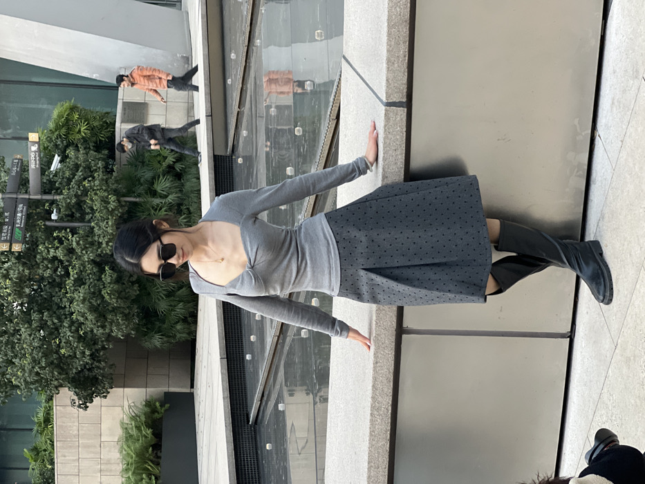 A female subject is positioned in an urban plaza in Yuzhong District, China. She wears a grey long-sleeved top with a knotted sweetheart neckline, a dark grey pleated midi skirt with subtle black polka dots, and black knee-high boots. Dark sunglasses cover her eyes, and she wears a gold-colored pendant necklace and a bracelet on her left wrist. She leans with both hands on a light grey concrete barrier. In the immediate foreground, to the lower left, a portion of a camera lens and dark clothing are visible, indicating a photographer. Behind the subject, a shallow rectangular water feature with faint reflections separates her from a paved pedestrian path. On this path, two males are walking from left to right; one wears dark attire and a face mask, the other an orange jacket. The background includes dense green foliage, a signpost with Chinese characters, and modern architectural structures composed of glass and concrete.