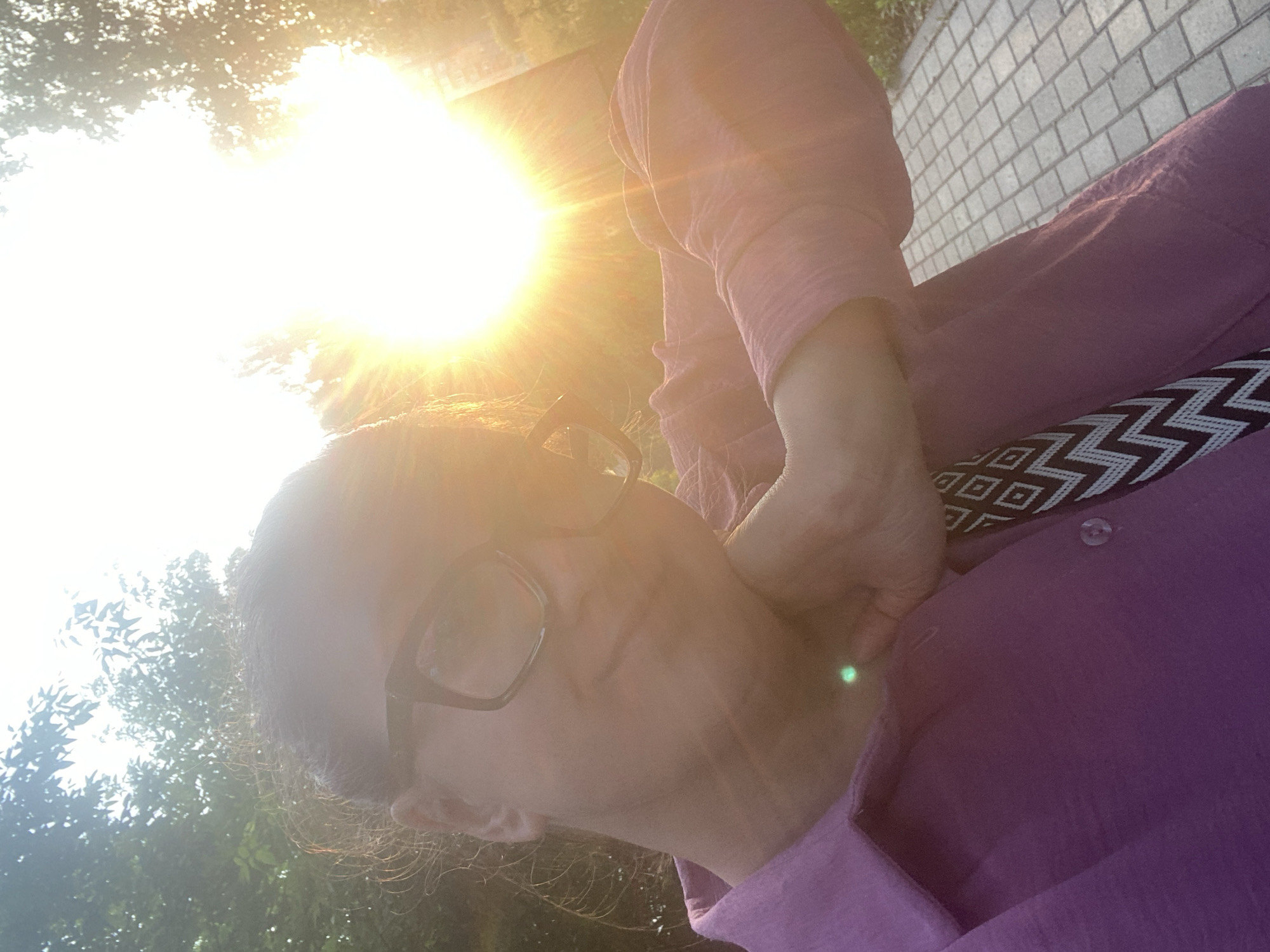 A woman wearing glasses and a purple collared shirt is taking a selfie outdoors during golden hour. The sun is setting behind her, creating a strong lens flare that dominates the upper right portion of the image. She has her right hand on her neck and her arm is bent. Her hair is pulled back. In the background, trees are visible on the left and a paved walkway with square stones is on the right, suggesting a park or a public outdoor area. The location is the city of Chaikovsky, Russia.