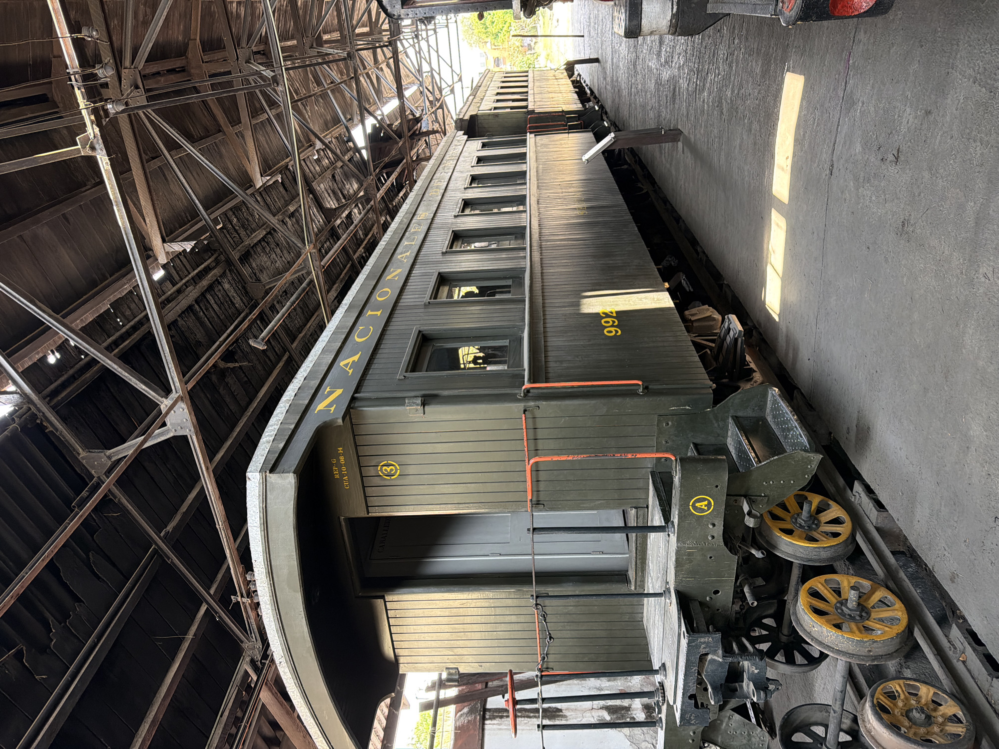 The image shows a vintage train car labeled "NACIONALES" with the number "992," housed in a wooden structure. The train car is olive green with yellow lettering and is partially under a covered area with a complex network of wooden beams and metal supports. The train car has a door and windows visible, along with steps leading up to the entrance. Train wheels are visible along the side. The setting appears to be a railway museum or maintenance facility. Although the location is stated as Cuautla, Mexico, the image itself lacks specific clues to confirm the location.