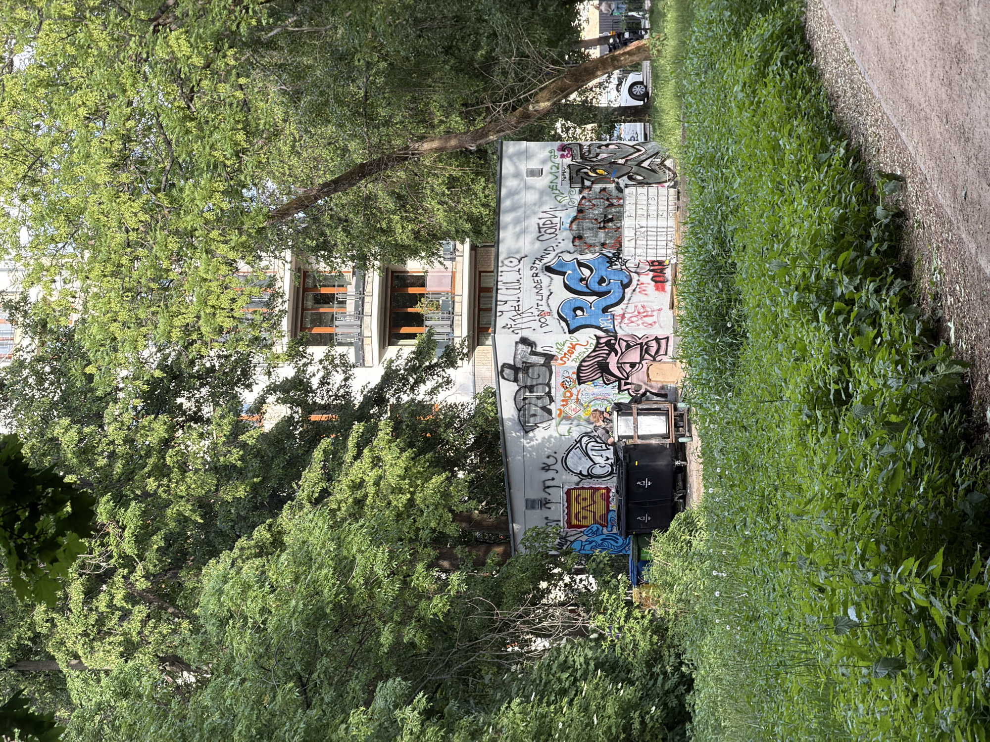 A sideways-oriented, medium shot depicts an outdoor scene featuring a building adorned with graffiti, located in Unknown, Unknown. To the left, the view is partially obscured by the dense foliage of several trees, some of which frame the building. The building, likely an apartment complex, showcases balconies with wooden railings.
A plain gray wall adjacent to the building is covered in various graffiti designs, displaying colors of blue, pink, gray, yellow, and black. Stacked on the ground in front of the graffiti wall are two black speakers and a small black television.
To the right of the wall, a dense patch of tall green plants extends to a sidewalk, with a clear separation between the grass and pavement. A section of the sidewalk appears in the frame.