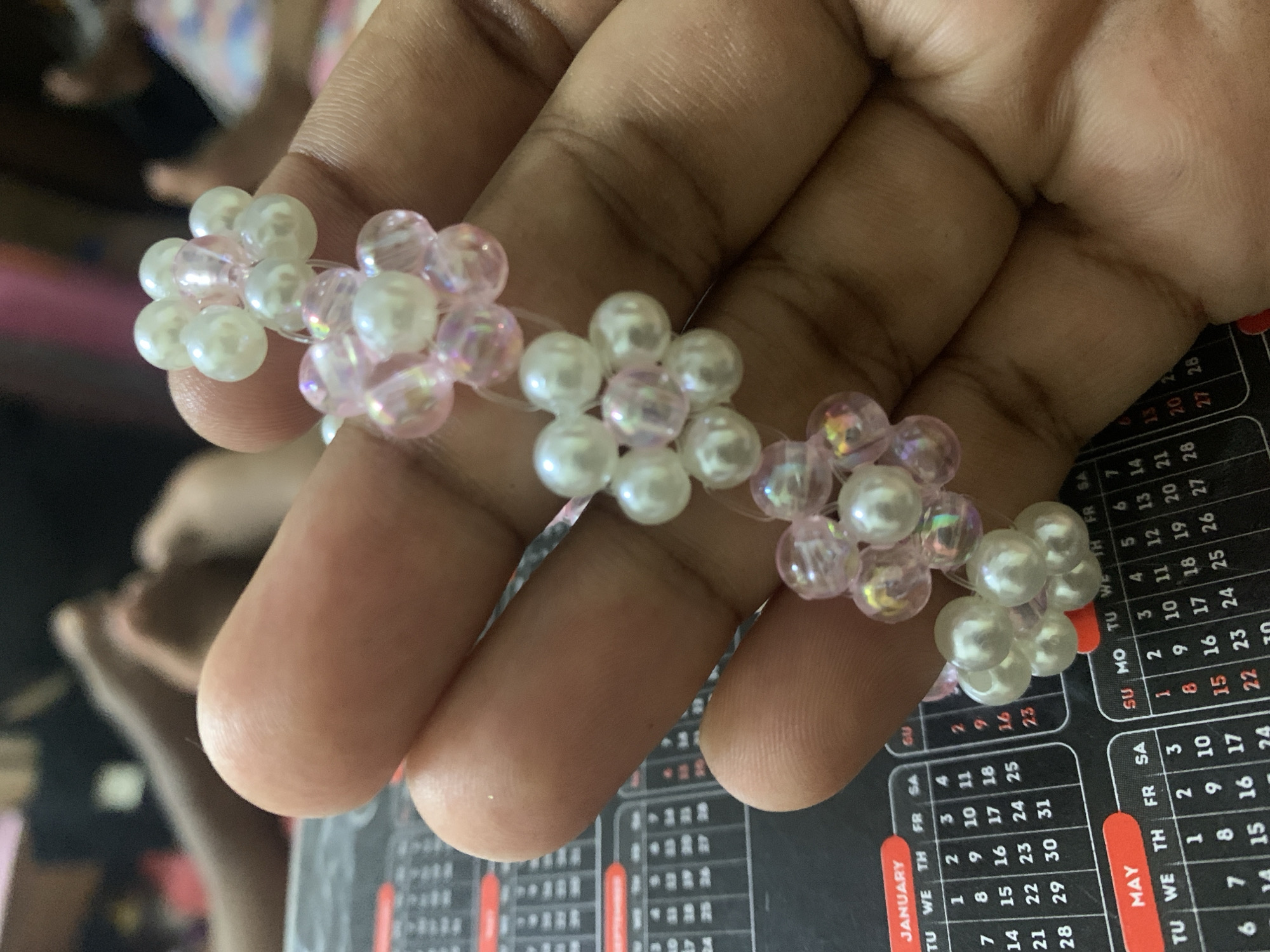 A hand is displaying a beaded flower ring made of white and iridescent pink beads. The ring is stretched across the fingers. The background is partially out of focus, but shows a desk calendar with dates and month names in English. There is no indication of Dar es-Salaam, Tanzania.