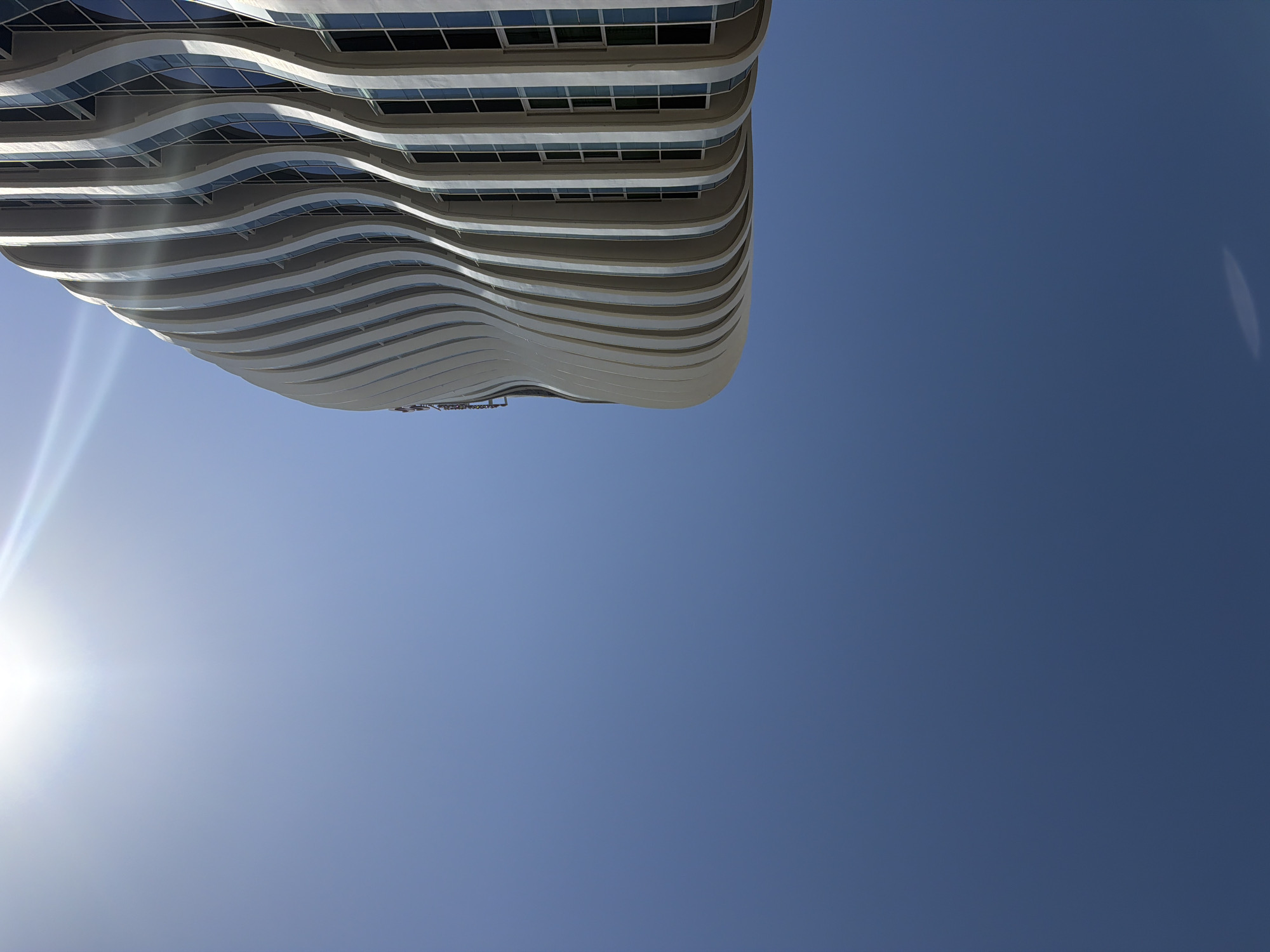 A low-angle shot captures the curved, white facade of a modern building against a clear blue sky. The building's exterior features a series of vertical, undulating fins that create a sense of fluid motion. Behind these fins, rectangular windows are visible, reflecting the bright sunlight. The sun, a powerful source of light, is positioned in the upper left corner of the frame, casting lens flares and bright highlights across the sky and onto the building's surface. The lower portion of the image is dominated by the vast expanse of the blue sky, with a subtle gradient from a lighter hue near the sun to a deeper blue at the bottom. The scene is devoid of any people or other immediate environmental details, focusing solely on the architectural form and the interplay of light and shadow.