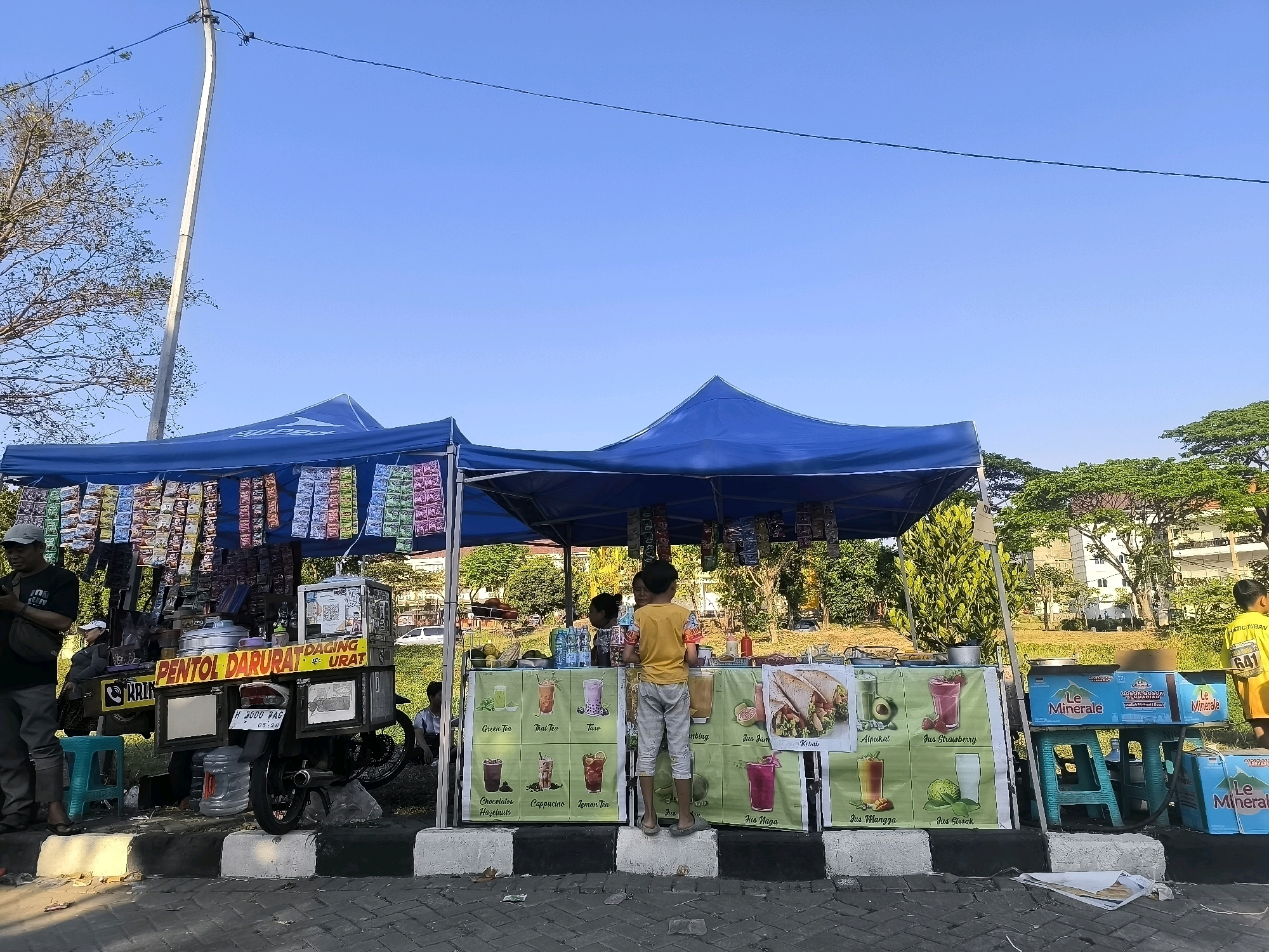 The image depicts a street food stall in Surabaya, Indonesia, on a clear day. The stall is set up under two blue tents. The left tent displays various packaged snacks hanging from above. The right tent advertises different beverages and food items with colorful posters featuring illustrations of drinks like "Green Tea," "Thai Tea," "Taro," "Chocolate Hazelnut," "Cappuccino," "Lemon Tea," "Avocado," "Strawberry Juice," "Mango Juice," and "Guava Juice," as well as "Kebab."
In front of the right tent, two young boys are interacting at the counter. One boy, wearing a yellow t-shirt and grey pants, is standing on a concrete block, looking towards the stall. Another boy, to his left, appears to be speaking with the vendor. A woman is partially visible behind the counter. To the far right, a person wearing a yellow shirt with the number "641" can be seen walking away.
To the left of the tents, a motorcycle is attached to a cart with "PENTOL DARURAT DAGING URAT" written on it, suggesting it sells pentol (meatball) dishes. A man in a dark shirt and grey pants stands to the left of this cart, looking at his phone. A blue stool is placed near the cart.
The stall is situated on a paved street with white and black painted concrete blocks marking the edge. In the background, there are trees and a building, indicating an urban environment. Several bottles of "Le Minerale" water are stacked on teal-colored plastic stools to the right of the beverage stall. The overall scene suggests a busy local market or street side where people gather to purchase food and drinks.