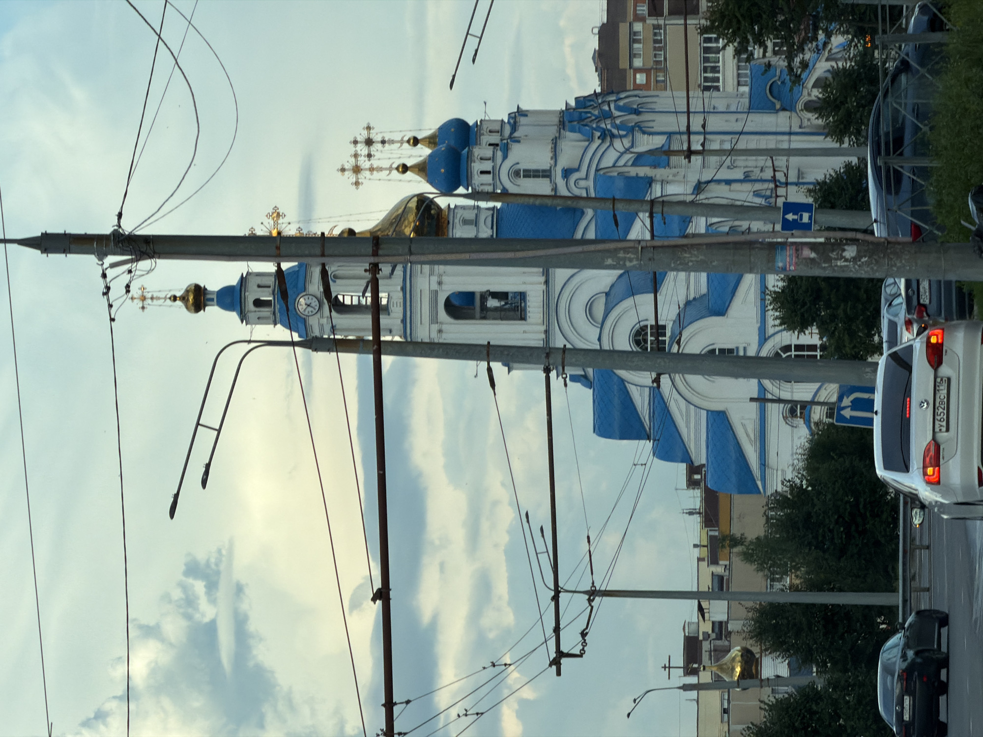 The image captures a view of the Kazan Temple in Kazan, Russia, from a street level perspective, likely from a moving vehicle. The temple, a prominent white and blue structure with golden domes and crosses, is partially obscured by overhead wires and utility poles, which dominate the foreground. Multiple streetlights are visible along the wires. The sky is overcast with scattered clouds, and the overall lighting suggests it is daytime. Several cars are present on the road, indicating an urban environment. In the background, some apartment buildings and trees can be seen. A traffic sign indicating a straight-ahead lane is also visible on a pole.