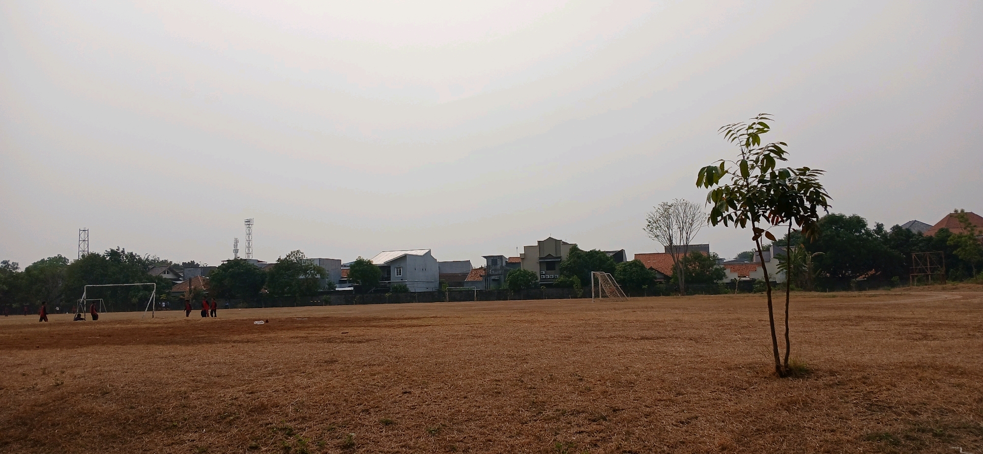 An outdoor shot of a dry, brown grass field with a sparse canopy of trees and buildings visible in the background. The sky is overcast. Several soccer goals are present on the field, and a group of children are playing soccer in the distance. A small tree stands in the foreground. The scene is likely a sports field in a residential area of Cirebon, Indonesia.