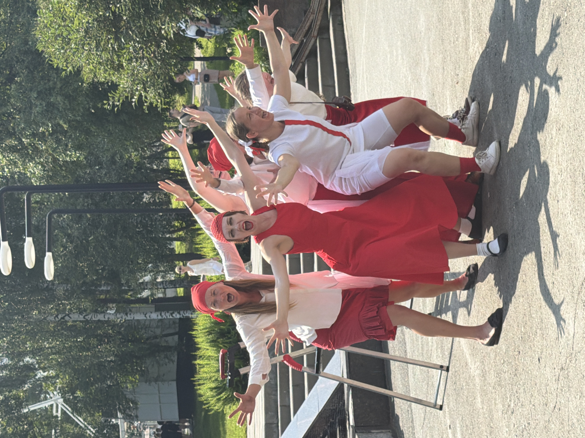 A group of six women are performing an outdoor dance or theater piece on a paved surface with trees and a building in the background. They are all wearing red headscarves and red and white costumes. The woman in the center foreground is wearing a red dress and has her mouth open in a yell. The woman to her left is wearing a white shirt and red skirt and is also yelling. The woman to the right of the woman in red is wearing a white top with red trim and white shorts, and she is also yelling with her arms outstretched. The other women are also stretching their arms and appear to be in motion. In the background, there are trees, a building, and what appears to be a set of stairs. The scene is lit by sunlight, casting shadows on the ground. This image was taken in Yekaterinburg, Russia, at the "Youth Palace" park.