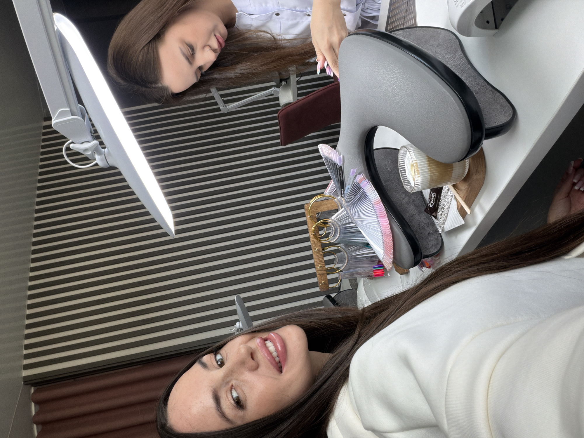 The image depicts a scene likely set in a nail salon or beauty studio in Izhevsk, Russia. Two women are present, viewed from an angled perspective as if taken from a selfie.
In the foreground, a woman with long dark hair and a light-colored top (possibly white) is smiling and looking directly at the camera. Her lips are glossy, and she appears to be taking a photo, capturing herself and the surrounding environment.
In the background, another woman with long dark hair is lying relaxed on a white table, with her eyes closed. She is wearing a white collared shirt. Her left hand, with long, light-colored nails, is visible near a grey, ergonomically shaped hand/wrist rest typically used for manicures. This suggests she is in the process of receiving a nail service or resting during one.
The white table is equipped with various items related to nail care. A bright, white LED desk lamp with an adjustable arm illuminates the area. Next to the hand rest, there are multiple fan-shaped display wheels showcasing a variety of nail polish colors. One of the swatches has "Liquid Peach #13" partially visible. A clear glass cup with a straw, containing a light-colored beverage (possibly a latte), sits on the table, alongside a small wooden stirring stick and a packet with the text "Kendal." A device, likely a UV/LED nail lamp, is partially visible in the upper right, with "SUNG" (possibly part of "SUNUV") visible on its surface. Under the table, a dark brown armrest or chair back is visible.
The background features a wall with horizontal stripes, alternating between dark grey/black and light grey/white. The overall setting suggests a professional and well-lit environment for beauty treatments.