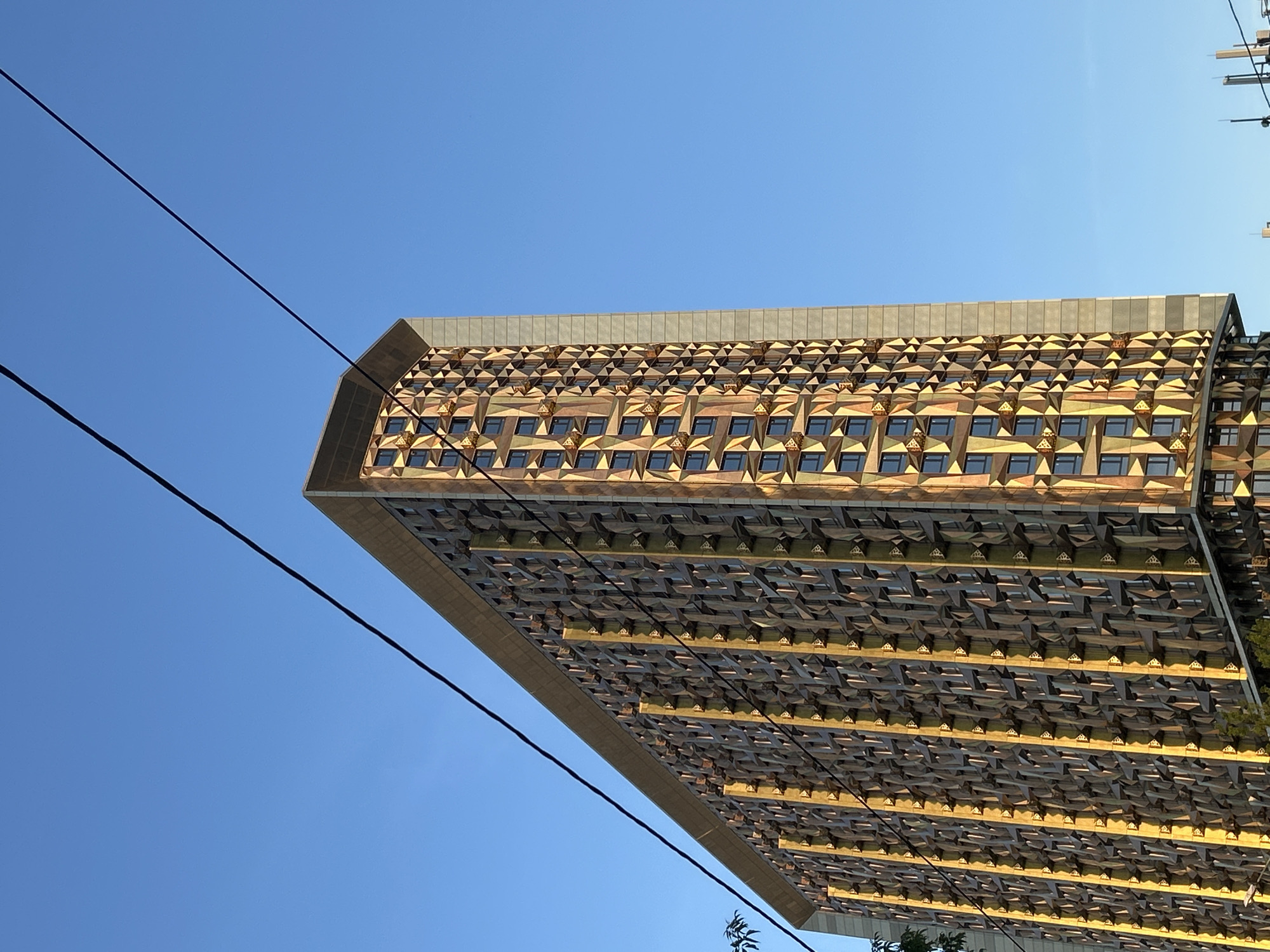 A large, modern building with a distinctive, intricately patterned facade is captured from a low angle against a clear, bright blue sky. The building's exterior is covered in a repeating geometric design, appearing as a tessellation of numerous small, triangular or pyramidal elements. These elements display a palette of gold, brown, and gray tones, with parts of the facade reflecting a warm, golden light, suggesting daytime illumination. Small, dark-paned windows are integrated into this complex pattern across multiple visible stories.
Two dark, thin lines, likely utility cables or power lines, traverse diagonally across the clear sky in the foreground, partially intersecting the building. The sky is cloudless, indicating fair weather. A small hint of green foliage is visible at the very bottom right of the image. No people or specific activities are depicted. The overall impression is one of modern architecture viewed under natural sunlight. No visible text is present.