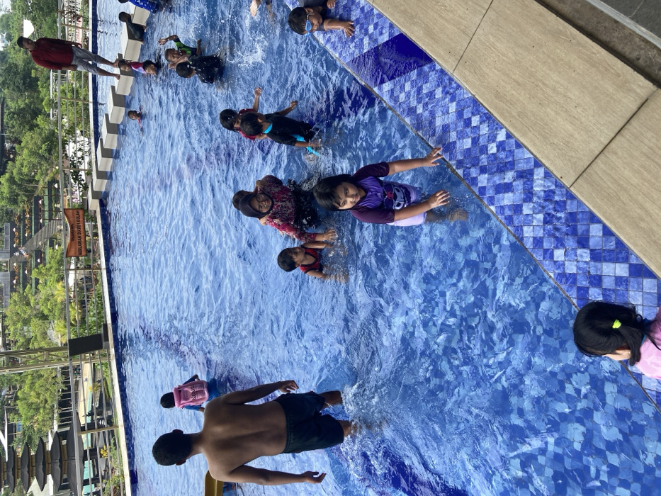 A group of people are in a swimming pool. The pool has blue water and a blue tiled edge. A shirtless man is standing in the water near the bottom of the frame. Several children are swimming, some are wearing swimsuits or rash guards. One woman in a floral shirt is surrounded by children. The pool is located outdoors with trees and buildings visible in the background.