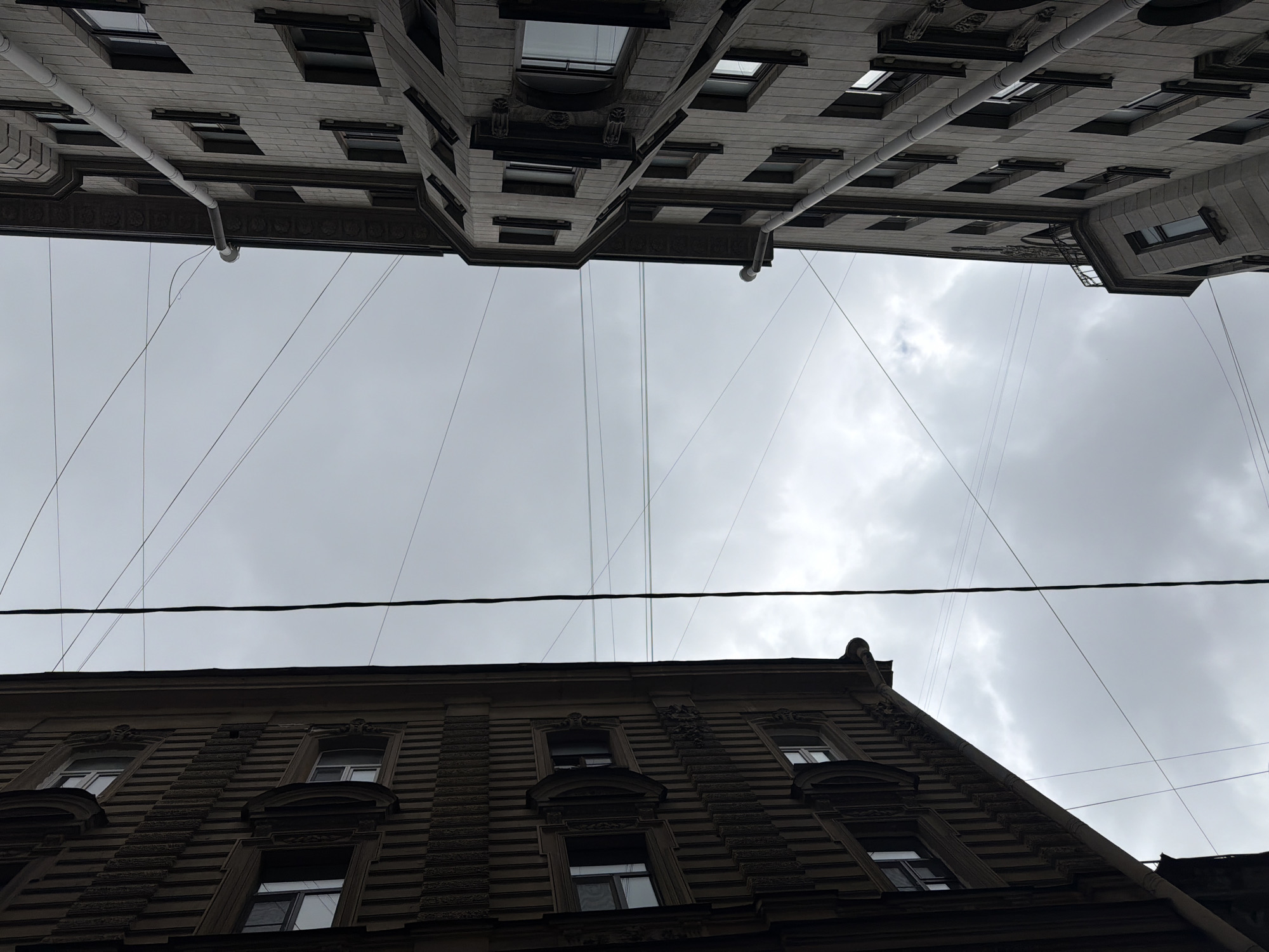 The image captures a view of two buildings in Saint Petersburg, Russia, seen from a low angle, looking upwards. The buildings are positioned on either side of the frame, with a cloudy sky visible between them. The building at the top appears to be a light-colored stone structure with multiple windows, and a drainage pipe is visible. The lower building is darker, with a textured facade and ornamented window frames. Wires stretch across the sky, connecting the two buildings. The scene conveys a sense of urban density and the architectural style common in Saint Petersburg.