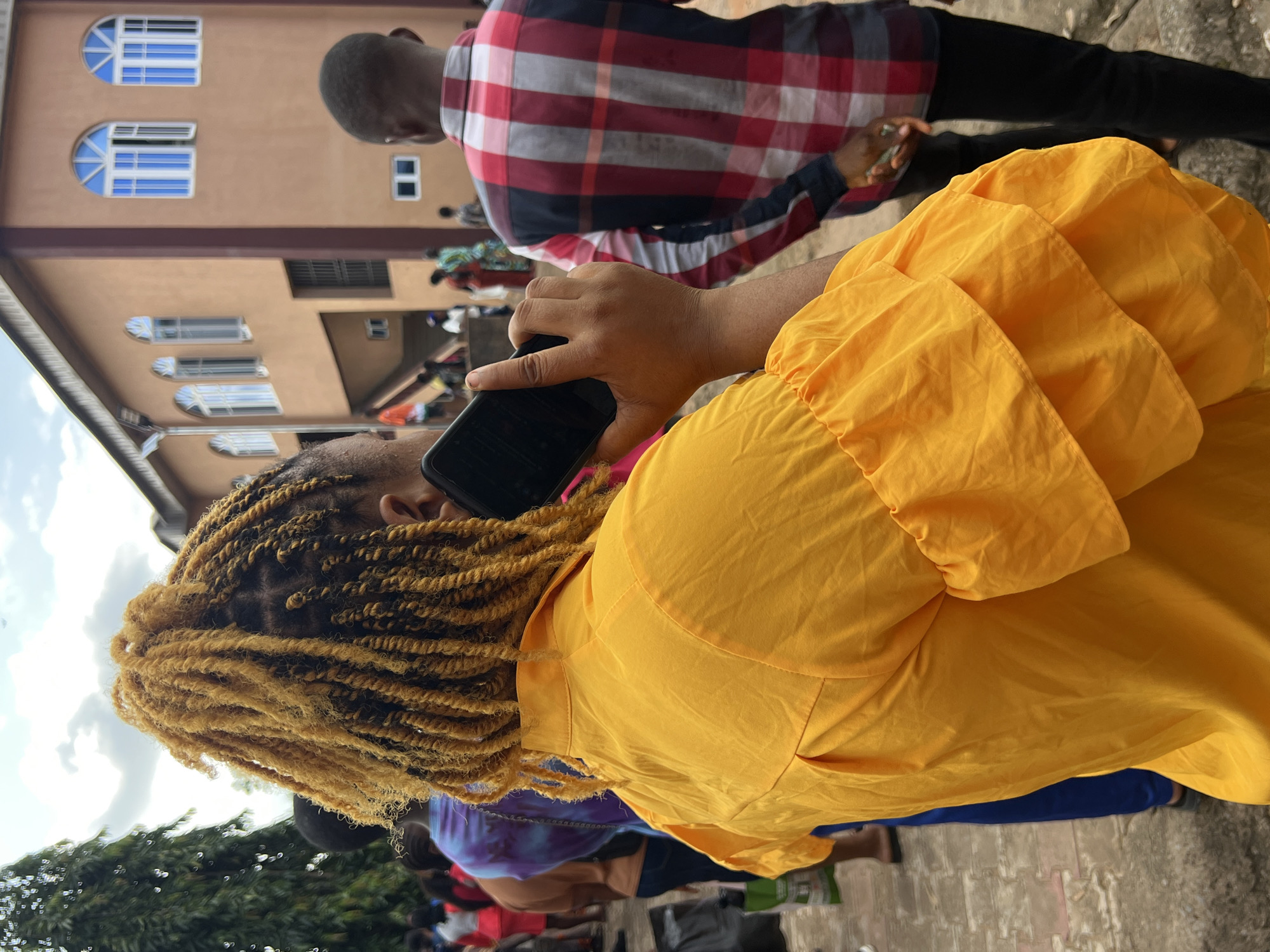 In Nnewi, Nigeria, a person with blonde-dyed braided hair holds a smartphone to their ear, standing in front of a building with a warm brown facade and arched blue-windowed openings. The person is wearing a vibrant yellow shirt with tiered, ruffled sleeves. To the right, another individual wearing a red, white, and grey plaid shirt and black pants stands with their back to the camera, holding what appears to be a small object in their hand. In the background, a line of people dressed in casual attire are visible, and a large green tree stands to the left of the frame. The ground is paved with bricks. The sky is overcast with clouds.