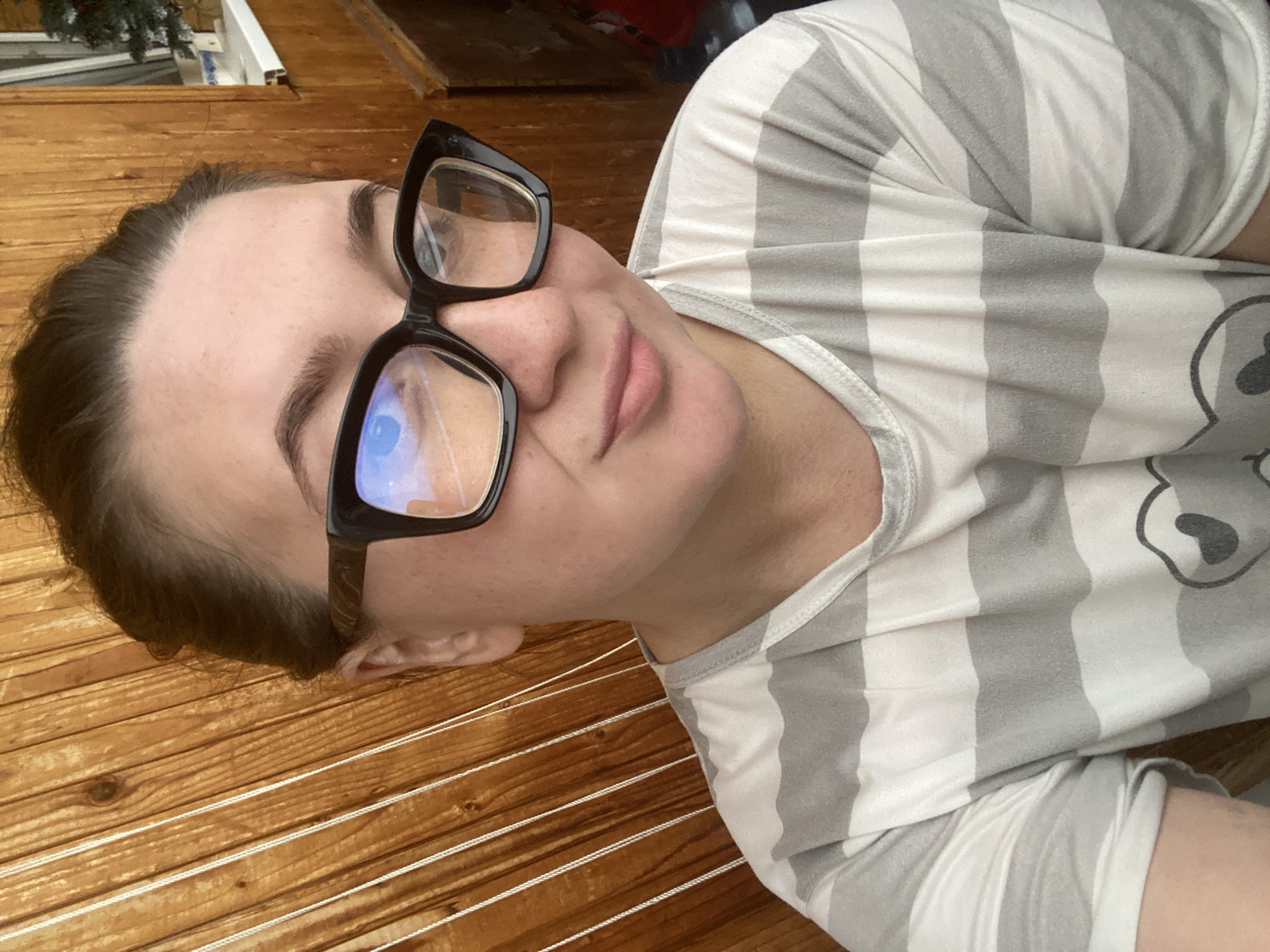 A woman with brown hair pulled back and wearing black-rimmed glasses is shown from the shoulders up in a selfie. She is looking slightly to her right and smiling gently. She is wearing a light gray and white striped t-shirt with a graphic print of a cartoon heart on it. The background is a light brown wooden wall, possibly an exterior wall or fence, with vertical planks. To the right of the woman, a white windowsill is visible, with a small, decorated evergreen tree on it, suggesting it might be near a window or door. A small portion of a red object is also visible in the far right background. The lighting suggests natural daylight. The location appears to be outdoors in a residential area in Chaikovsky, Russia.