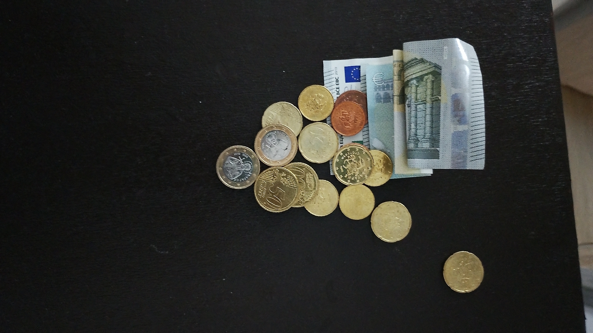 The image displays a collection of Euro banknotes and coins scattered on a dark, textured surface, possibly a table. The banknotes are partially visible, showing architectural imagery and the Euro symbol, suggesting they are of higher denominations. The coins, varying in size and color, are arranged in a cluster and individually. Notably, there are gold-colored coins, some with visible markings, and a few reddish-brown coins. One coin, larger and bi-metallic, is also present. The lighting casts subtle shadows, highlighting the texture of the surface and the metallic sheen of the coins. The background is a dark, solid color with minimal detail, keeping the focus on the currency. There are no people, animals, or clear indications of specific actions beyond the placement of the items. The scene appears to be a still life composition of money. There is no information or visual cue in the image that suggests a location in Smolyan, Bulgaria, or any specific city.