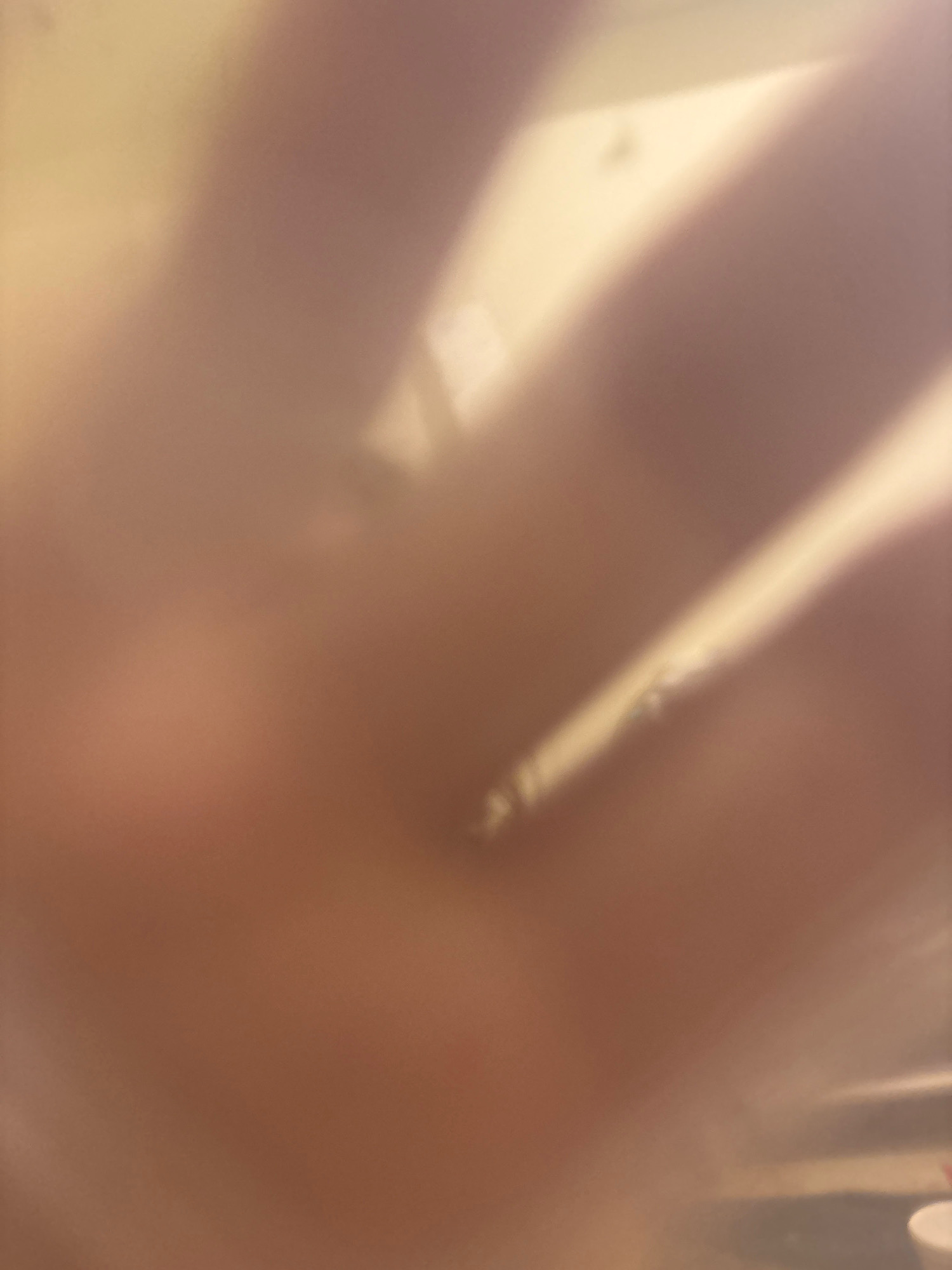 The image shows a close-up, out-of-focus view of a hand, possibly held against a frosted glass or plastic surface. A bright light source casts a diagonal glow across the hand, highlighting some of its contours. The background is a soft, diffused blur of light and shadow, with no discernible details of the surrounding environment, including any specific location in Ashvem, India. The image is primarily composed of warm, muted tones of beige, brown, and pale yellow. There are no identifiable objects or actions beyond the presence of the hand and the lighting.
