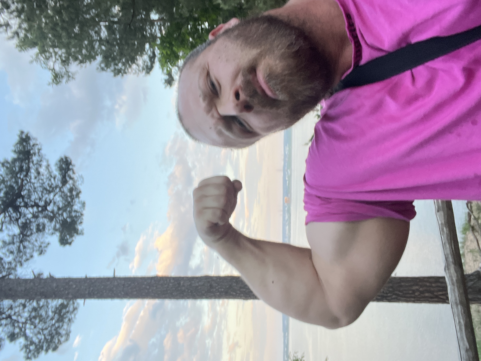 A man with a beard is posing with his arm flexed in an outdoor scene. He is wearing a pink shirt with a black strap across his chest. The background shows a cloudy sky, trees, and what appears to be a body of water. A tree trunk or branch is visible in the middle of the frame. The image appears to be taken from a low angle and is oriented upside down. It is impossible to confirm the location of Noviy, Russia, in this image.