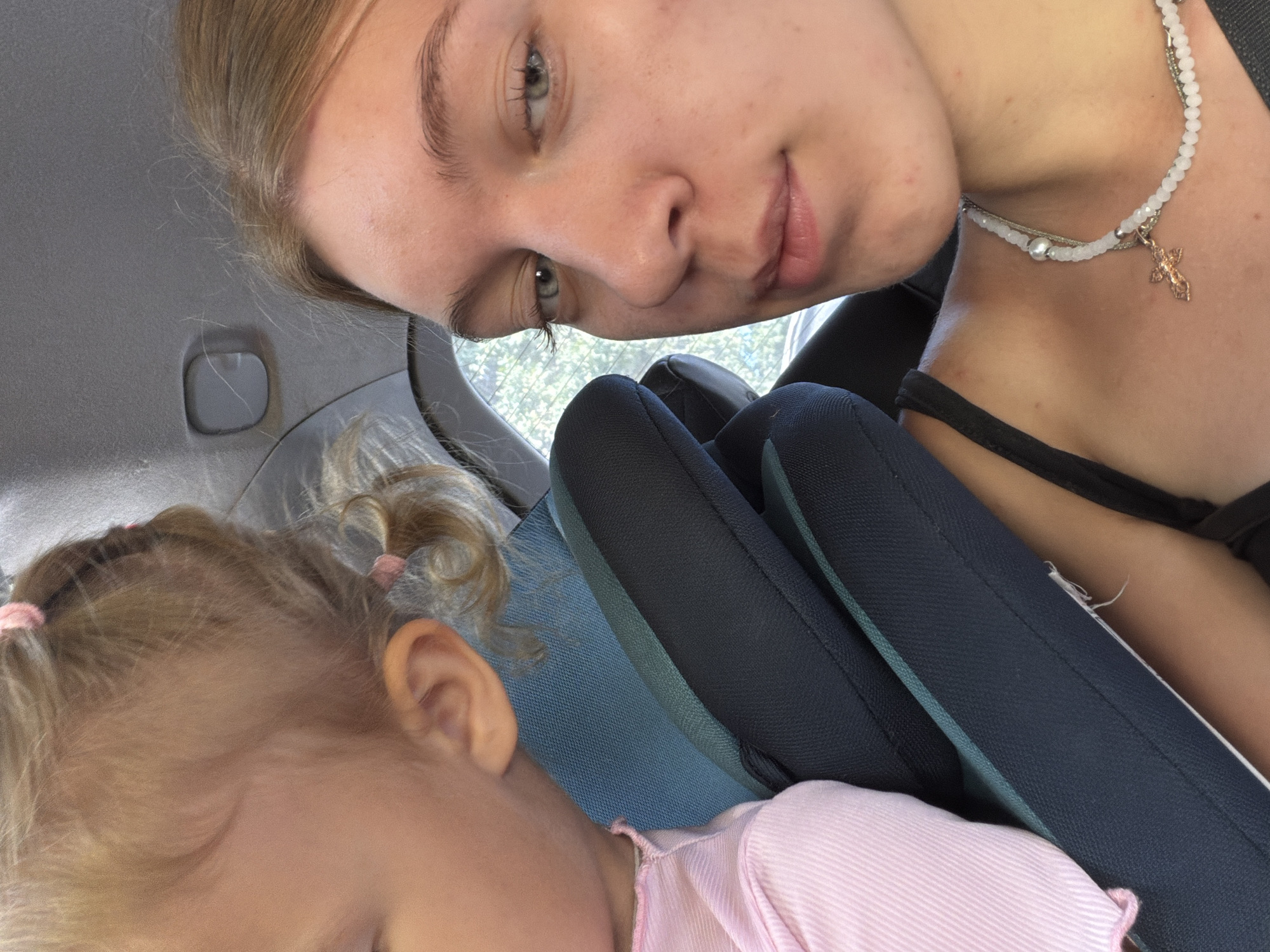 A young woman and a toddler are seated in a car. The toddler, on the left, has blonde hair styled in pigtails with pink elastics and is wearing a pink shirt. The woman, on the right, has blonde hair and is wearing a black tank top and a white beaded necklace with a cross pendant. They are both positioned in car seats. The interior of the car is visible, including the headrest and ceiling. The background shows blurred greenery through the car window, suggesting an outdoor environment. The scene appears to be a casual moment captured inside a vehicle, likely during a trip.