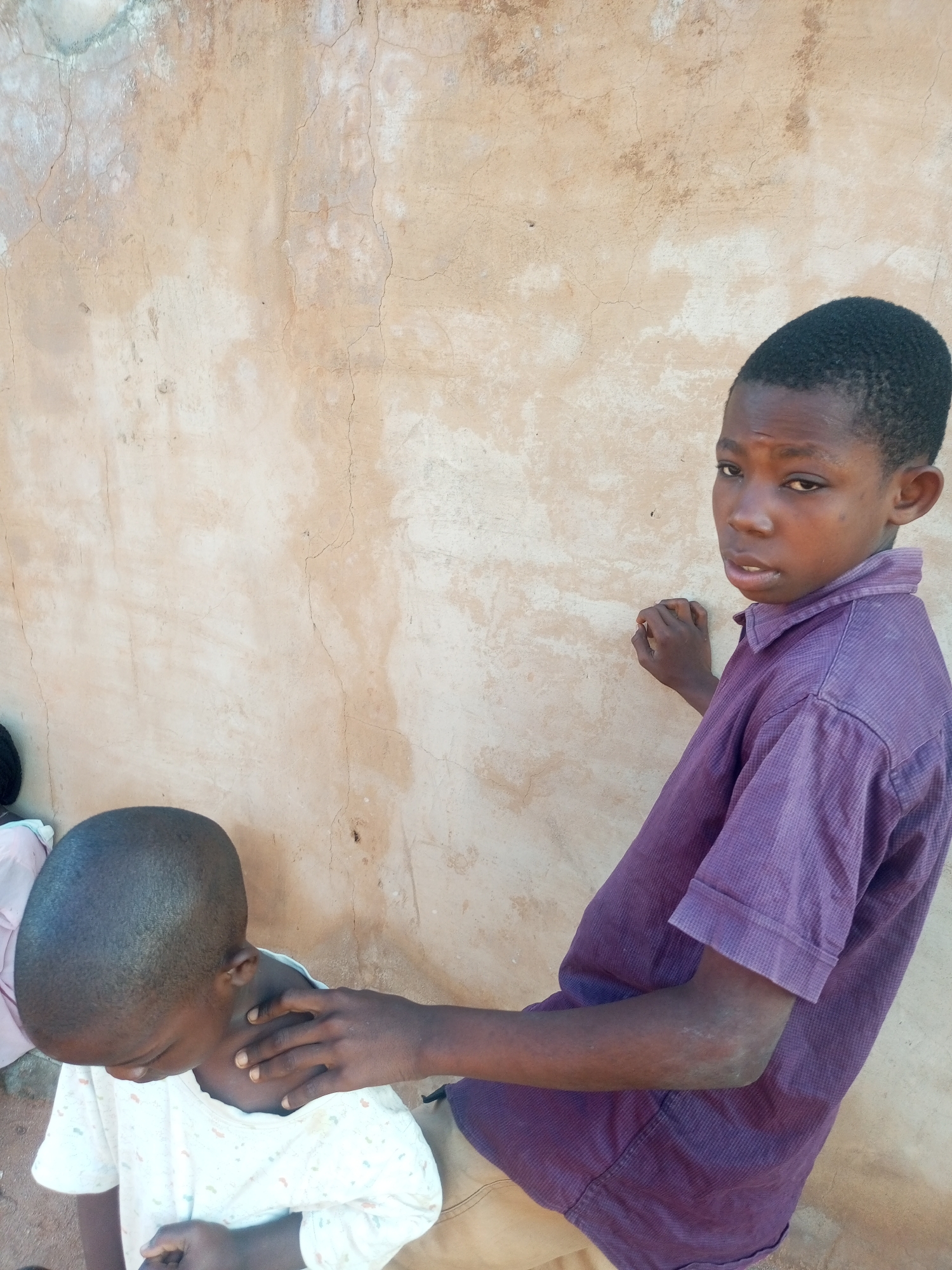 In Tempure, Nigeria, two young boys are seen standing near a textured, light-colored wall. The boy on the right, wearing a purple, collared shirt, is looking towards the camera with a concerned expression. His right hand is resting on the shoulder of the boy on the left. The boy on the left, facing away from the camera and looking downwards, is wearing a white shirt with a subtle pattern. His head is shaved. The wall behind them exhibits cracks and variations in its surface.