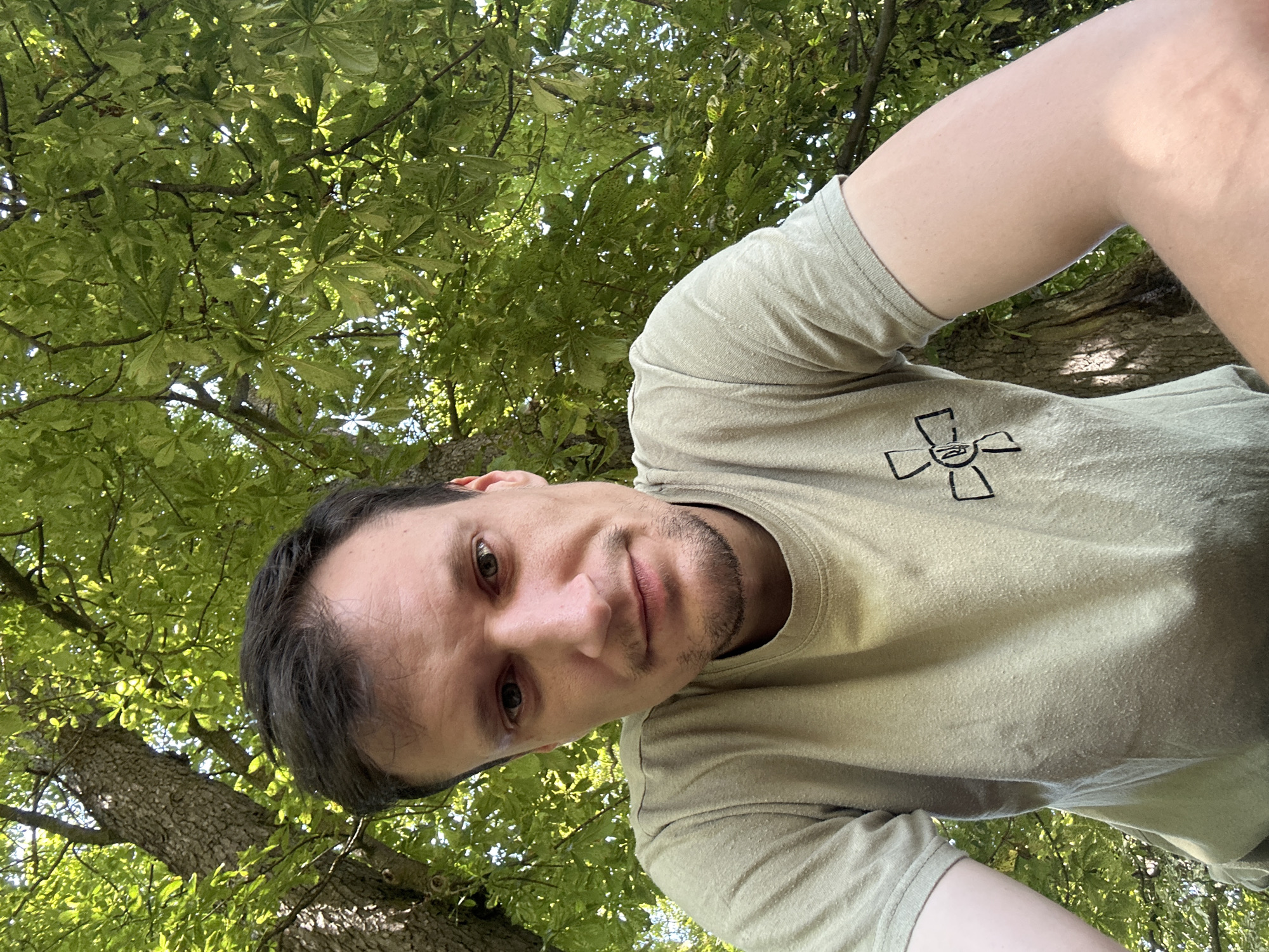 A man is looking directly into the camera, with a tree and lush green foliage behind him. He has short, dark hair and stubble. He is wearing a light green t-shirt with a black symbol on the left chest. The symbol is a circle with four outward-pointing arms, each divided into two sections, with a central circle. The man appears to be outdoors, with sunlight filtering through the leaves. The setting is identified as Makiv, Ukraine.