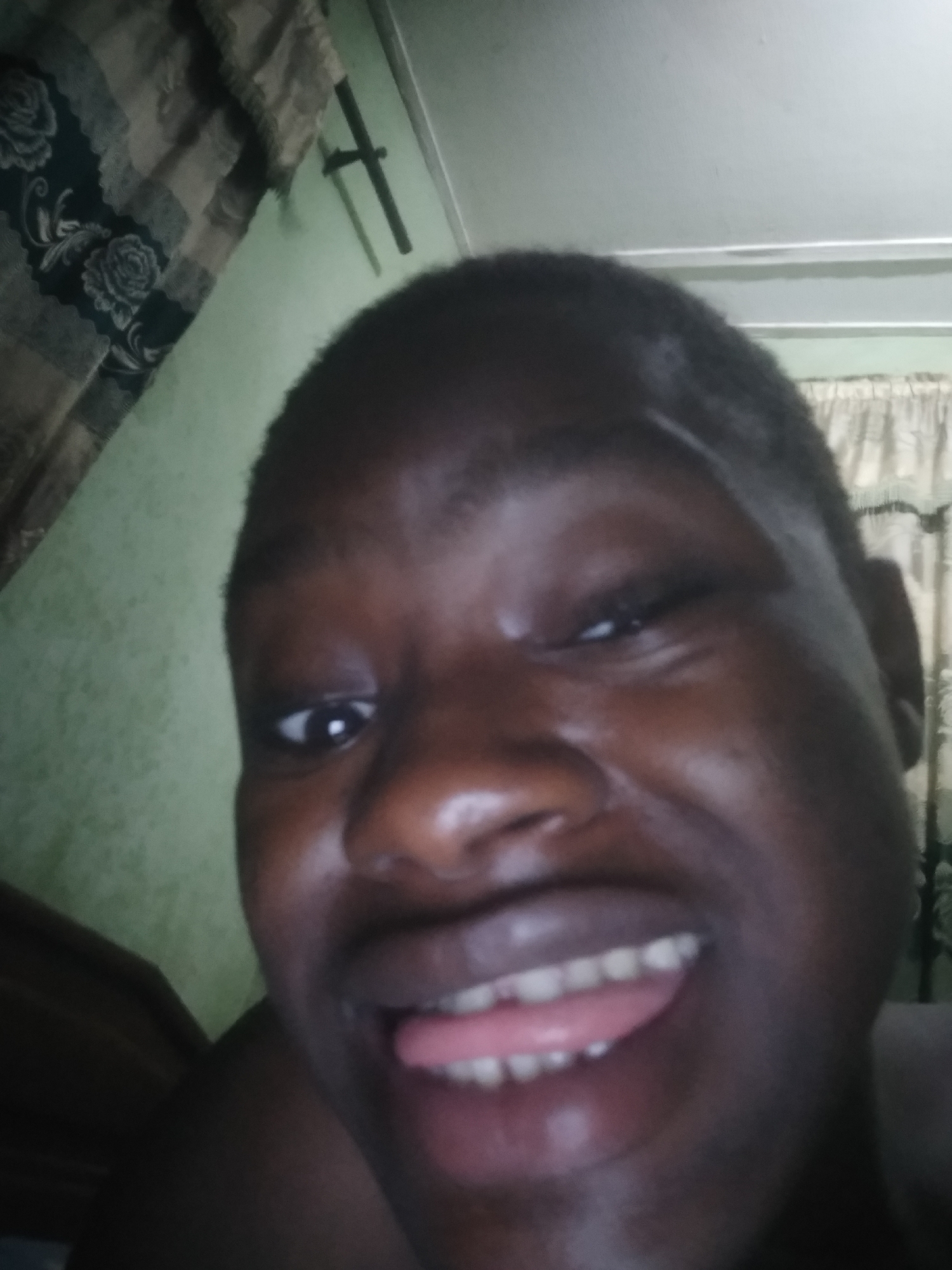 A close-up selfie of a person with dark skin, protruding their tongue, and smiling. The background shows a wall and curtains with a rose pattern. There is no definitive evidence to place the location specifically in Shangisha, Nigeria, from the image alone.