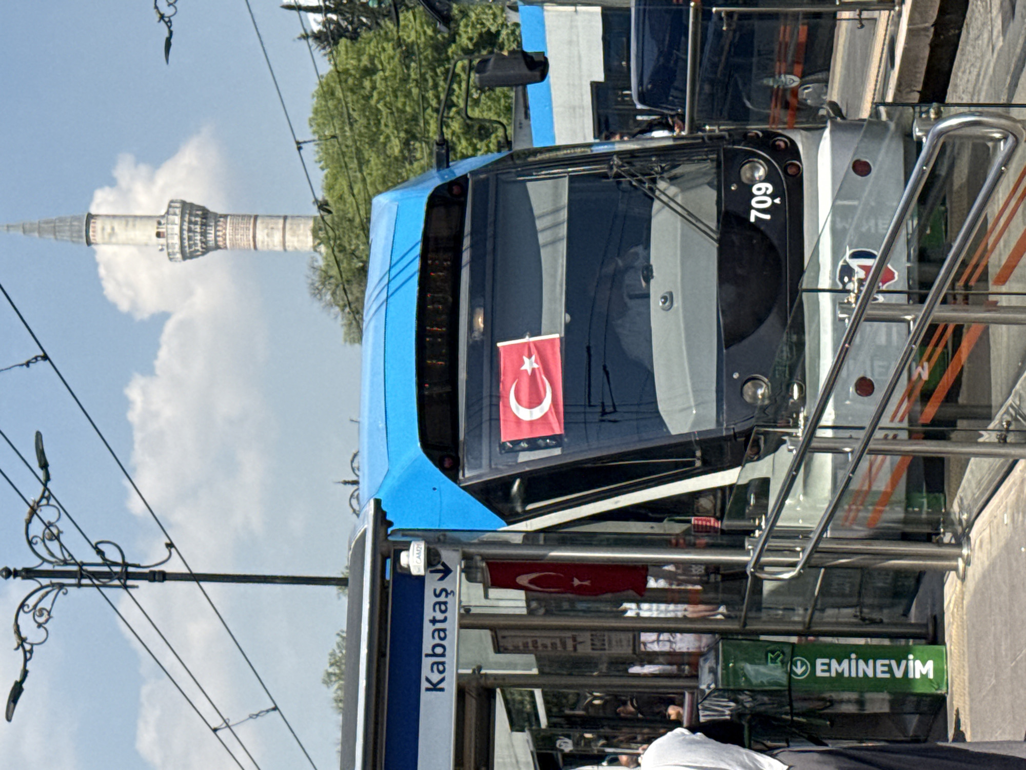 Here is a detailed description of the image:
The image captures a modern tram in the city of Istanbul, Turkey, featuring the distinct blue and silver color scheme. The tram is adorned with Turkish flags displayed in its front windows and along the side. The number "709" is clearly visible on the front of the tram. The tram station is marked with signs indicating "Kabataş" and "Eminevim," suggesting the tram's route or stops. The backdrop of the tram includes an ornate minaret rising against a blue sky with white clouds, giving a sense of the city's historical architecture. In the foreground, passengers can be seen waiting, and a few decorative lampposts with ornate designs add to the streetscape.