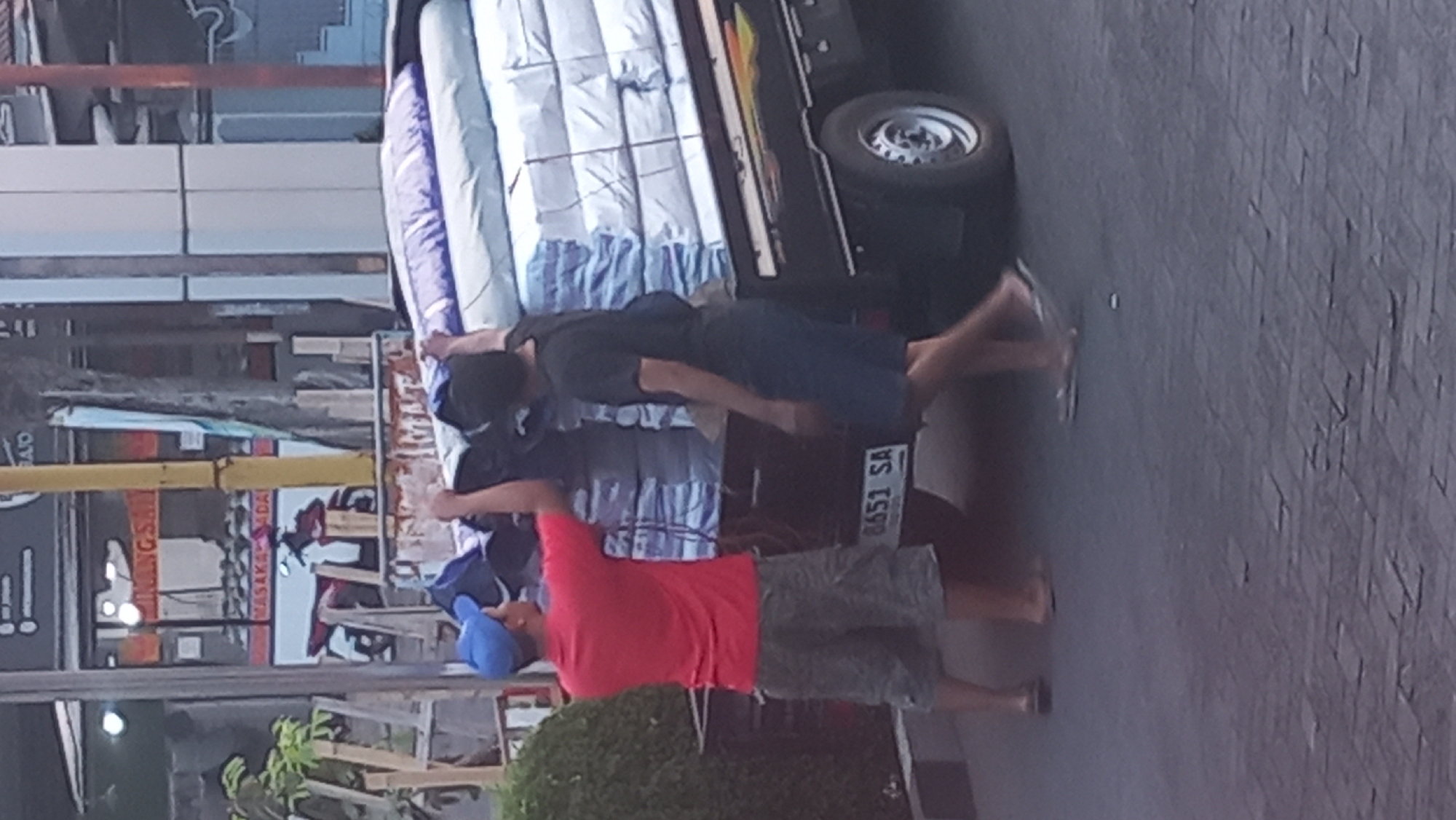Two men are loading large, rectangular packages onto the back of a dark-colored pickup truck in what appears to be an urban or semi-urban environment. The packages are stacked high and are covered with a blue tarp or plastic sheeting. One man, wearing a red t-shirt, blue cap, and patterned shorts, is on the left side of the truck, facing away from the camera and appears to be adjusting the load. The second man, wearing a black t-shirt and dark shorts, is on the right side of the truck, also facing away from the camera, and is reaching to help with the packages. The truck has a license plate that reads "8651 SA". In the background, there are buildings with signage, some trees, and a yellow pole, suggesting a commercial or street-side location. The ground is paved with what looks like interlocking bricks. The lighting suggests it is either early morning or late afternoon.