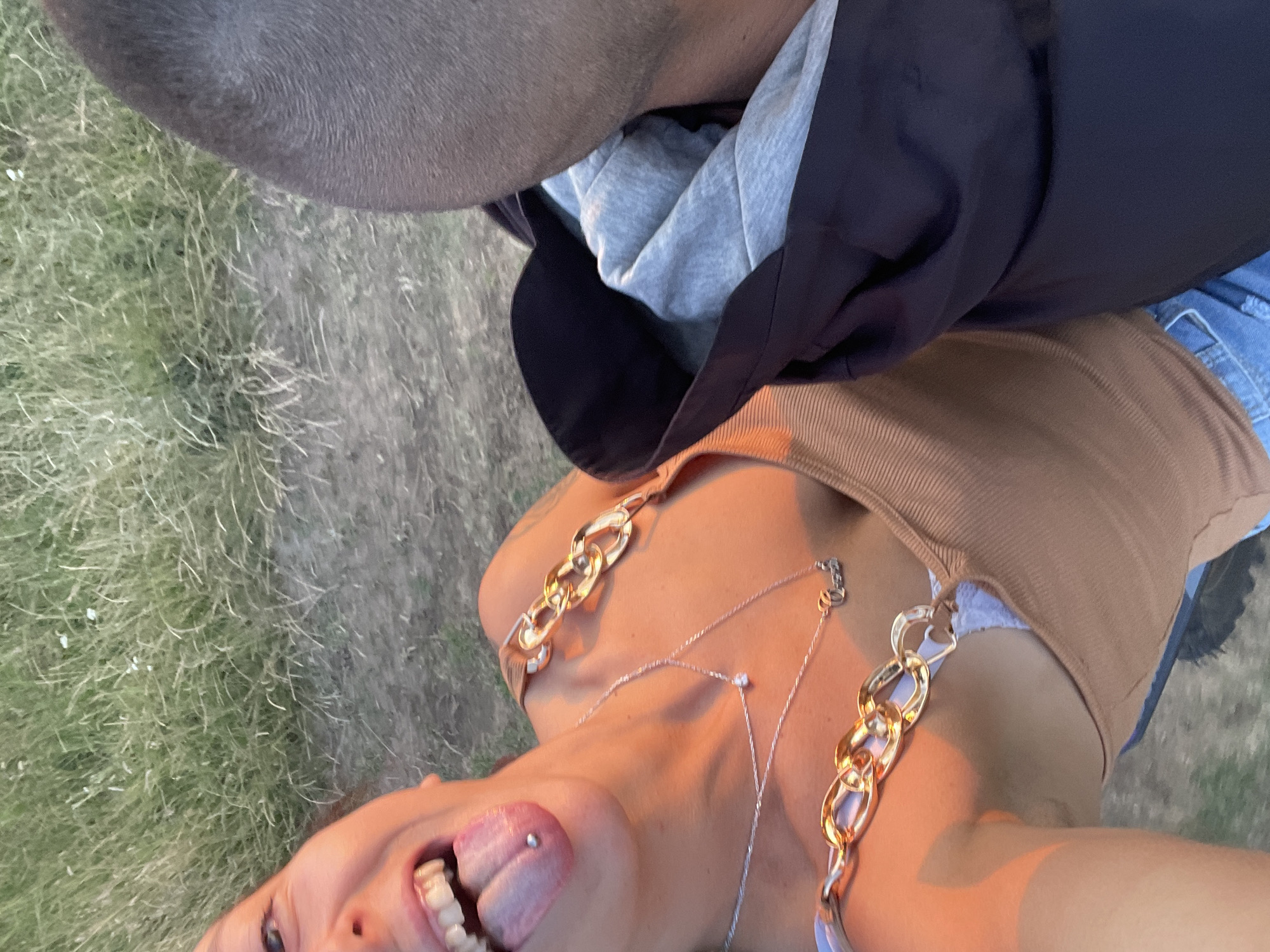 In an outdoor setting with dry, light green grass in the background, a young woman is shown from the chest up, taking a selfie. She has her tongue out, revealing a tongue piercing. She is wearing a tan, ribbed tank top with decorative gold chain straps. A silver necklace with a small diamond pendant and another chain with a word pendant hang around her neck. The image is cropped to include the back of a man's head on the right side of the frame. The lighting suggests it is either sunrise or sunset, casting a warm, orange glow on the woman. The location is stated to be Zheglartsi, Bulgaria.