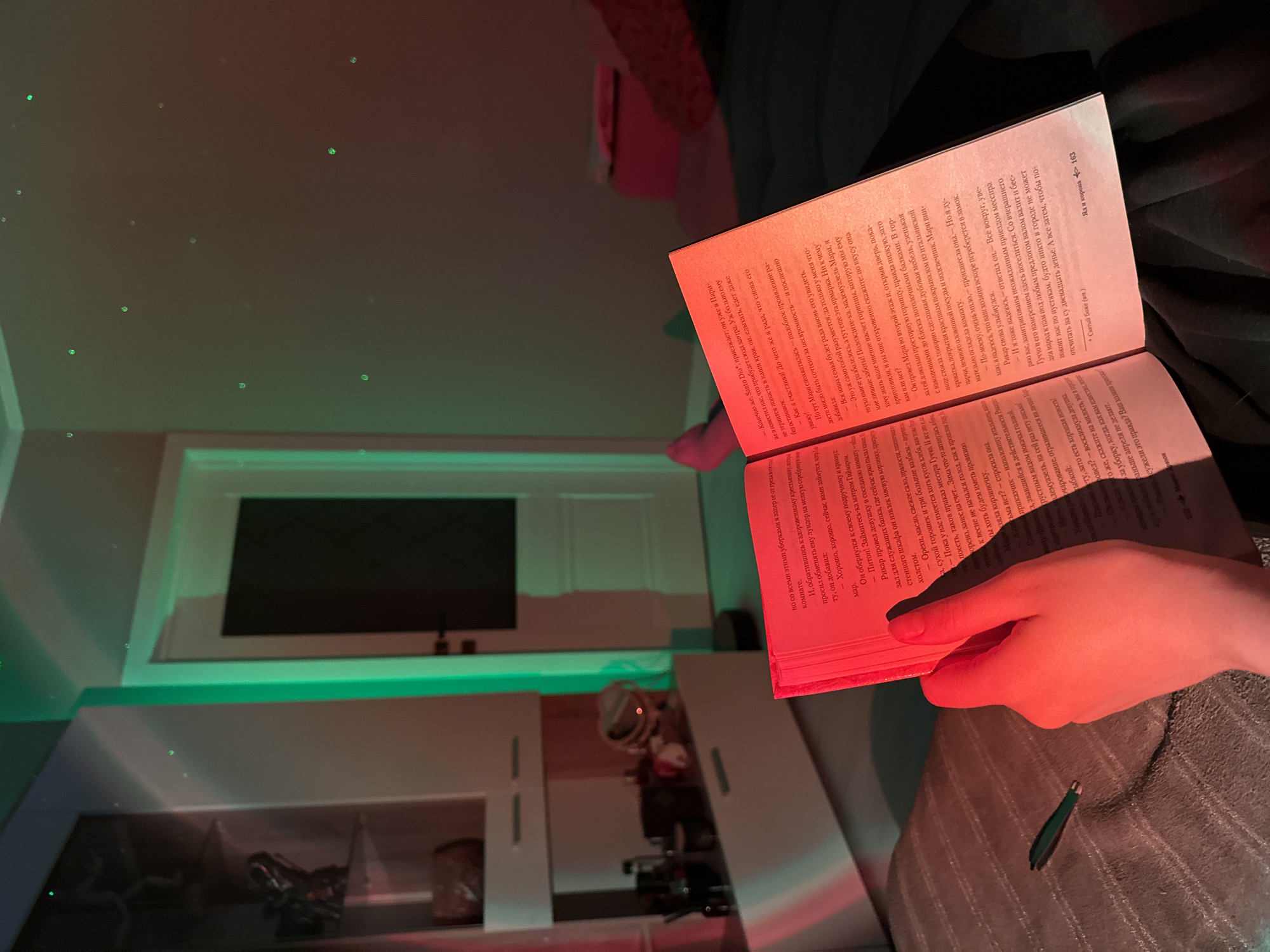 Here is a detailed description of the image:
**Scene:** The image captures an indoor scene with a person reading a book. The environment is somewhat dimly lit, with a play of colored lights – predominantly red and green.
**People:** There is a person's hand visible, holding the book open. The hand appears to be turning the pages, suggesting the person is actively reading.
**Subjects:** The primary subject is the book. The pages are filled with text.
**Objects:**
* **Book:** An open book with printed text.
* **Furnishings:** A sofa with a grey striped pattern is under the book.
* **Console Table:** It is in the background with visible items on the shelves.
* **Tweezers:** A pair of tweezers lie on the gray material, likely the sofa.
**Actions:** The main action is reading. The person is holding and manipulating the book, presumably to read its contents.
**Interactions:** The primary interaction is between the person and the book. The person is engaging with the text.
**Environment:** The environment appears to be a room with a modern aesthetic, based on the console table and lighting. There are some lights and spots on the walls, which create a pattern. The colored lights suggest a relaxed ambiance.