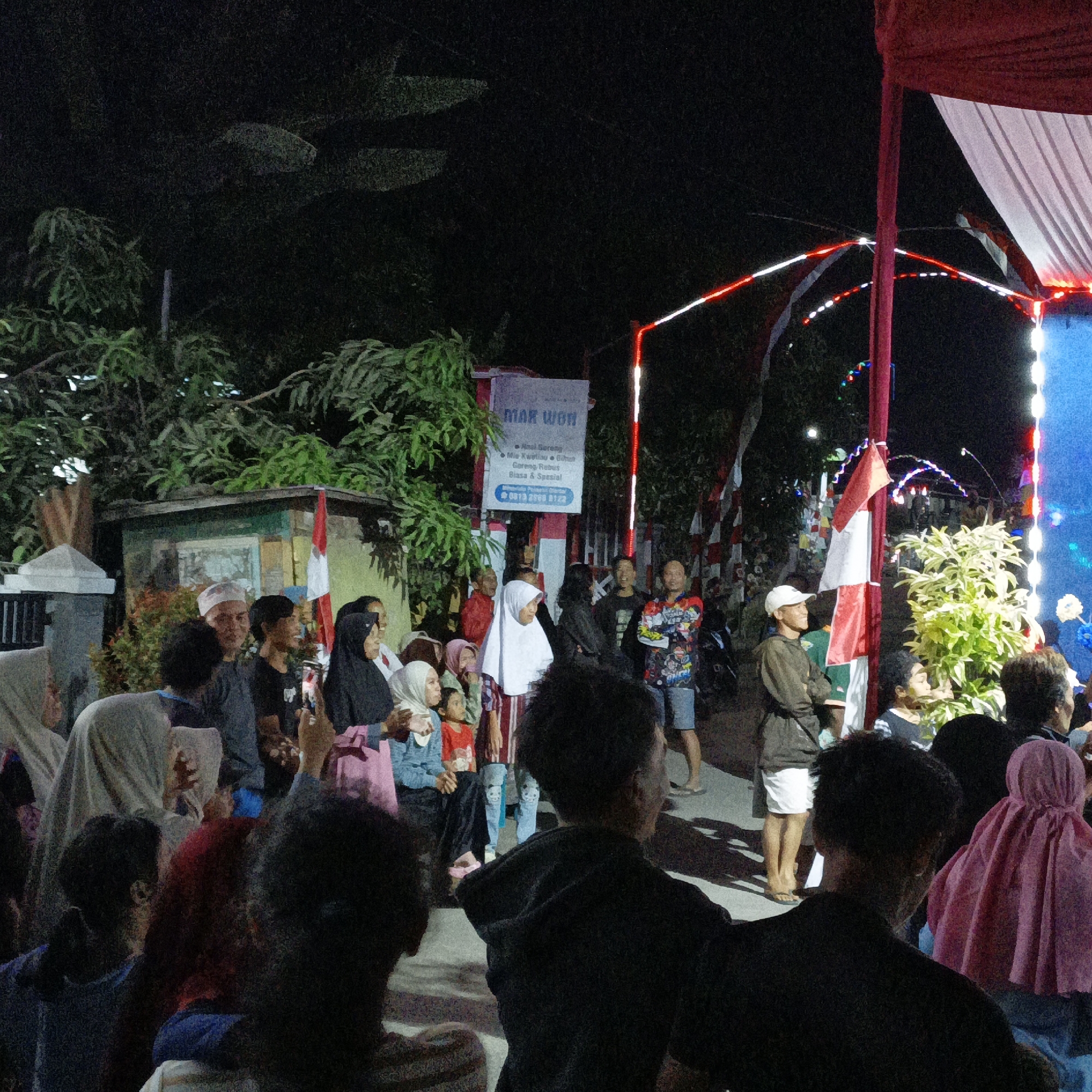 The image depicts a nighttime gathering of people in what appears to be an outdoor area in Tanahbaya, Indonesia. A red and white tent structure is prominent in the right side of the frame, decorated with red and white lights. In the background, trees and foliage are visible. Several individuals are present, some are looking towards a central point, possibly an event or performance, which is partially obscured. Some people are holding what appear to be phones, suggesting they might be recording or taking pictures. A sign with the text "MAR WON" is visible on the left side of the image, indicating a possible business or establishment. The overall atmosphere seems festive and social.