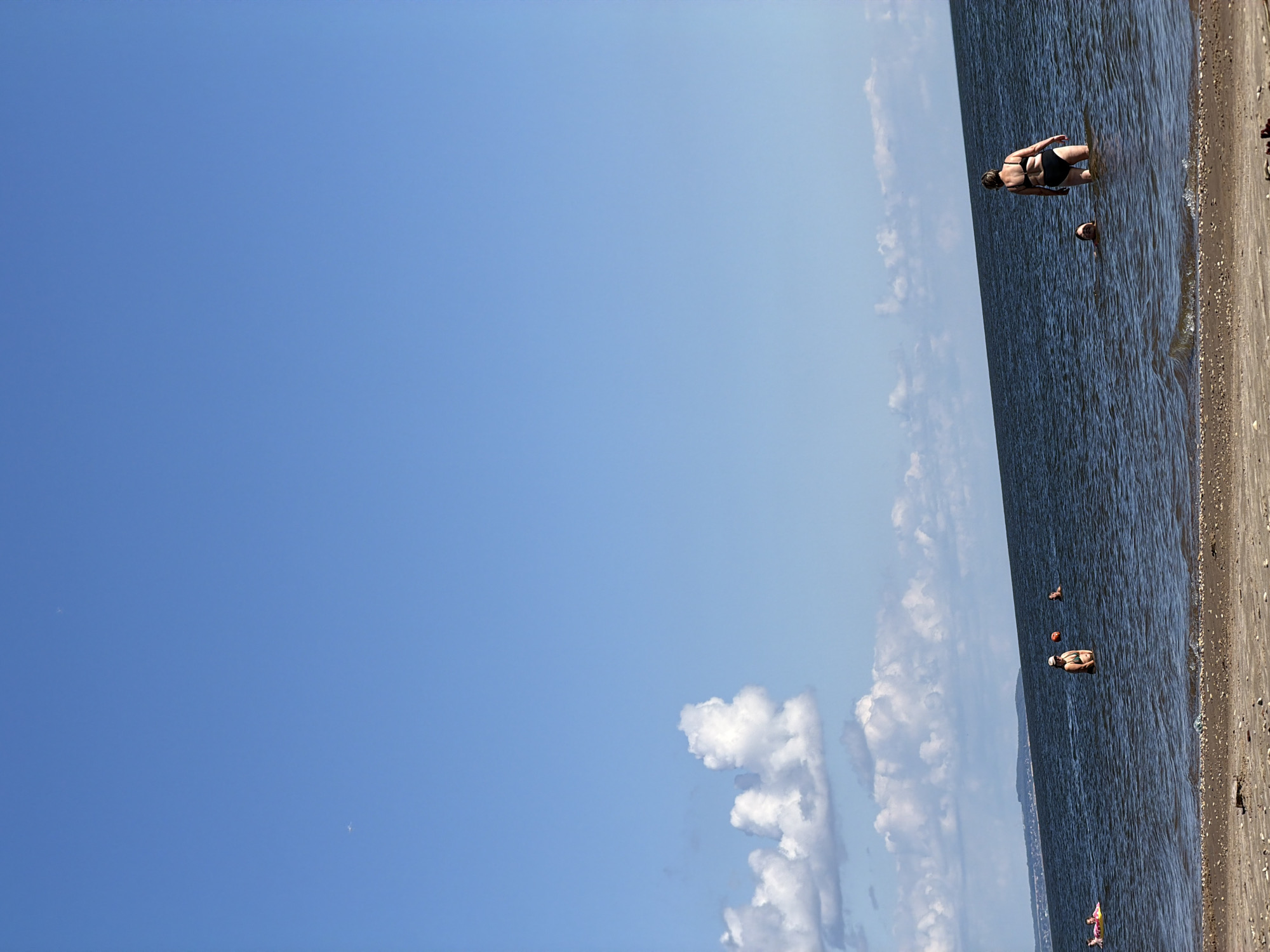 The image depicts a sunny day at a beach. The sky is a vibrant blue with a few scattered white clouds. The sea is calm and deep blue, with gentle waves lapping at the shore. The beach itself is made of sand and pebbles.
In the water, several people are enjoying the day. On the left, a person is on an inflatable toy. Further into the water, another person is visible. In the middle of the frame, a woman in a black bikini stands in the water, with an orange ball near her. Another person is swimming nearby. Towards the right, a woman in a black bikini is partially submerged in the water.
In the distance, a coastline with hills can be seen under the horizon. The overall scene is one of a peaceful and enjoyable day at the beach.
