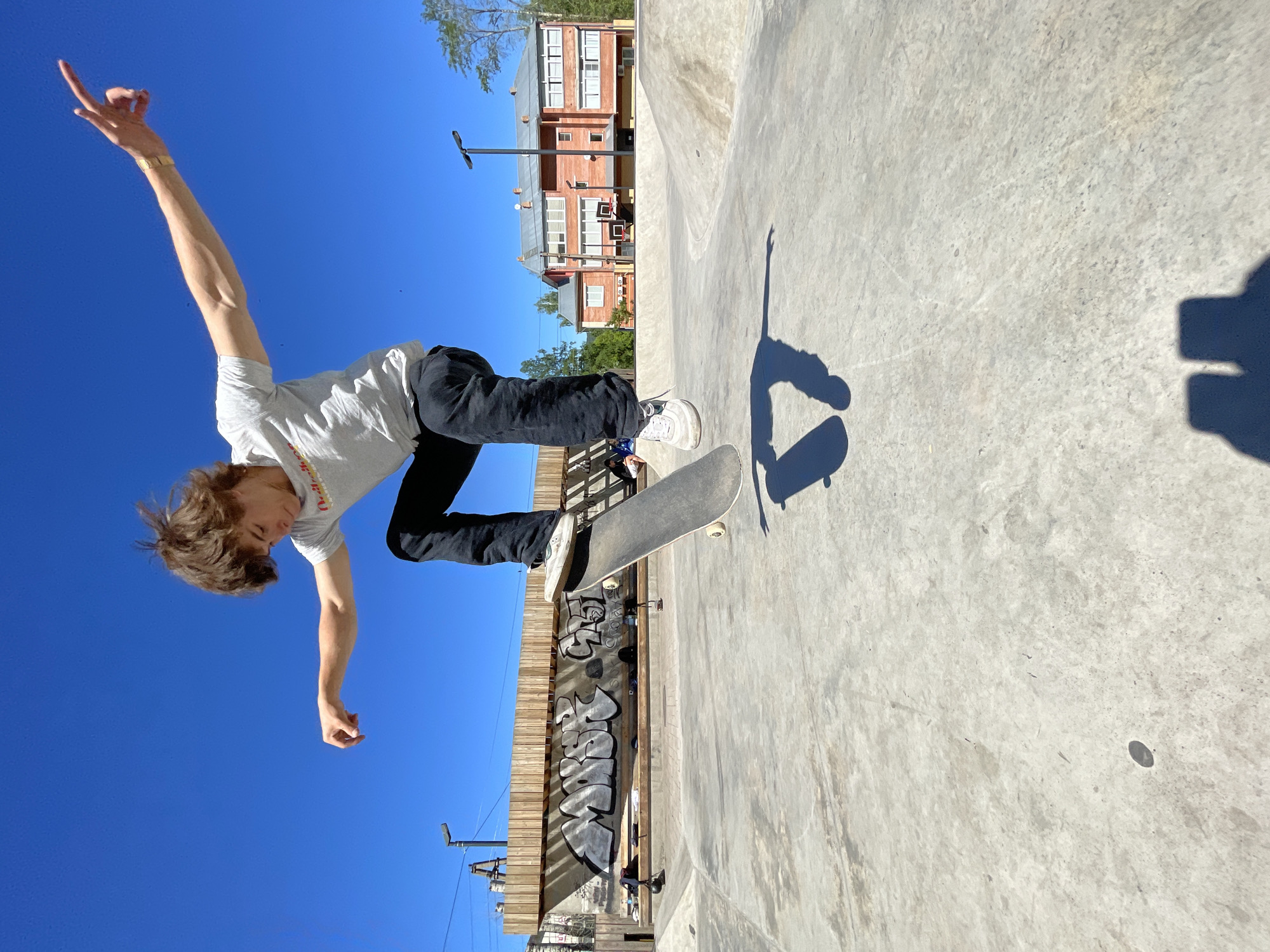 An adolescent male with light brown hair is captured mid-air performing a skateboarding trick against a clear blue sky. He is wearing a light gray t-shirt with a red logo and dark pants. His skateboard is angled beneath him, with the front wheels lifted off the ground. His arms are outstretched to his sides for balance, and his gaze is directed downwards. The scene is set in a concrete skatepark, with ramps and graffiti art visible in the background. The skatepark appears to be located in Divnogorsk, Russia, as indicated by the style of architecture and the presence of a red brick building with white-trimmed windows in the distance. Sunlight casts a distinct shadow of the skateboarder on the concrete surface below.
