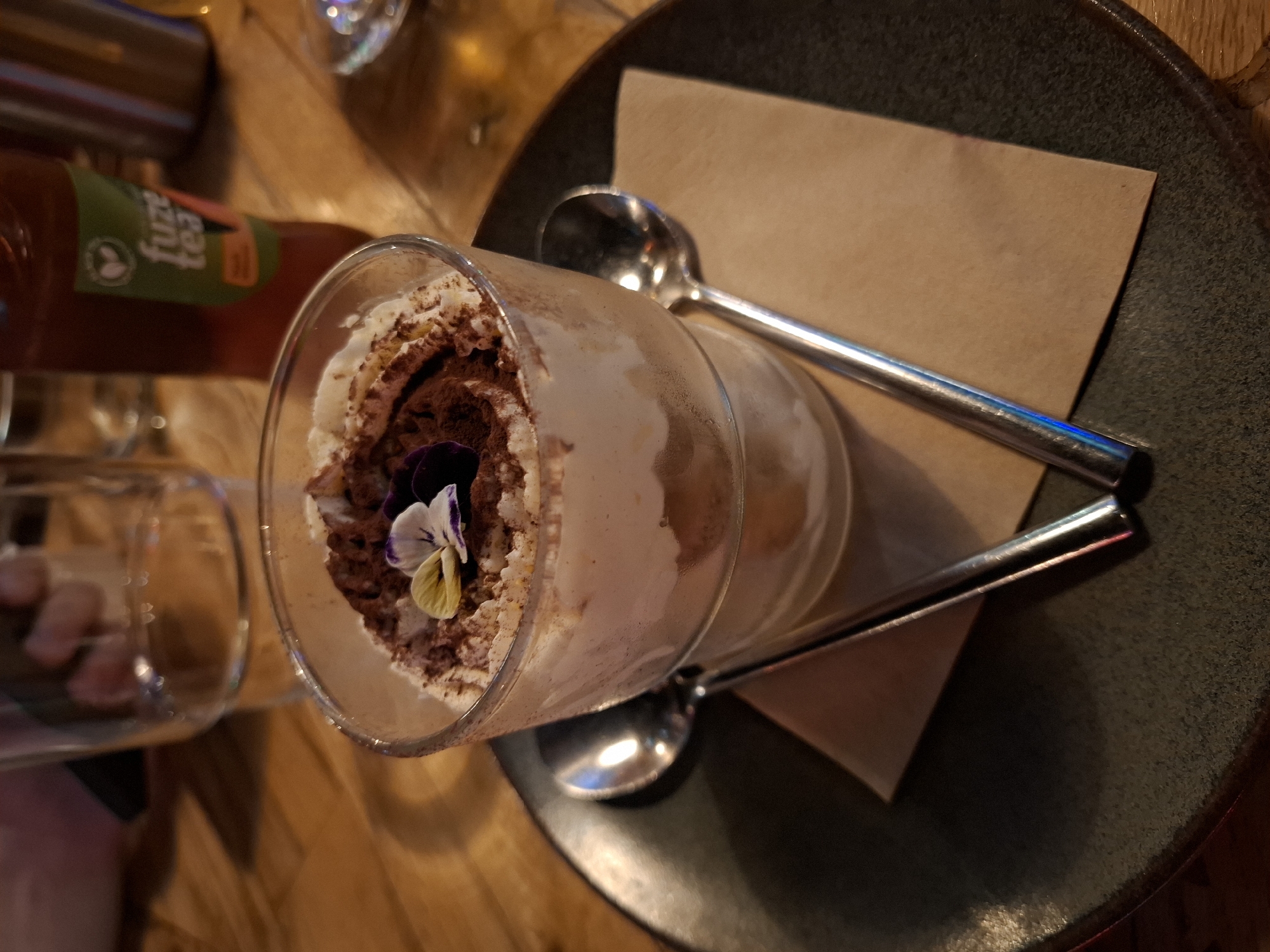 A close-up, eye-level shot shows a dessert in a clear glass bowl, topped with whipped cream and cocoa powder, and garnished with two small edible flowers. Two silver spoons are placed on a brown napkin, which is on a dark plate. In the background, a glass of water and a bottle of Fuze Tea are visible on a wooden table.