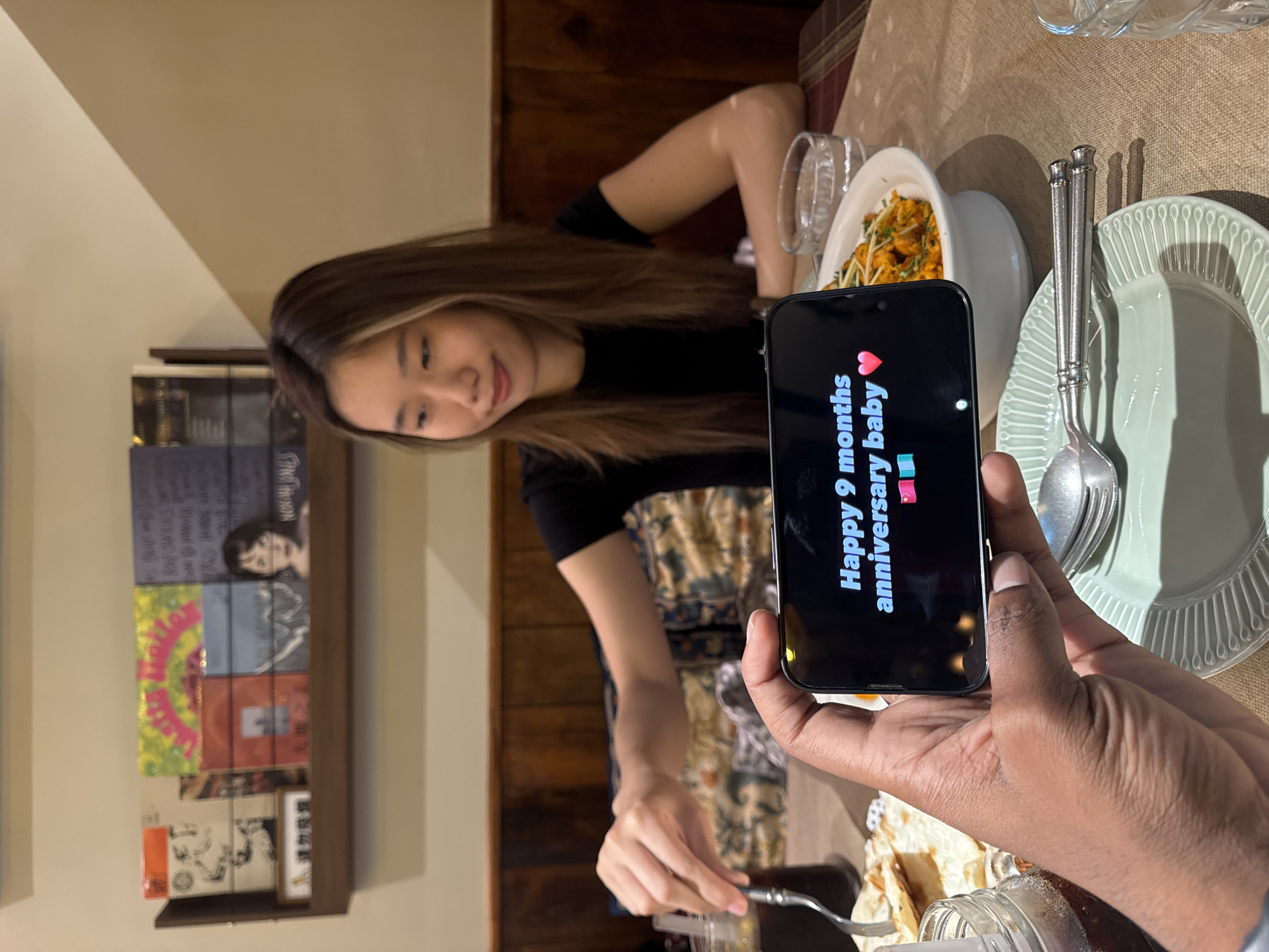 The image shows a woman at a table in an indoor environment, possibly a restaurant. She is facing towards the camera, smiling subtly. In front of her is a plate with food, and she appears to be using cutlery. Another person is holding a phone in the foreground, displaying the message "Happy 9 months anniversary baby" with heart and party popper emojis. Dishes, silverware, and possibly other food items are also visible on the table. The environment includes a shelf with what appears to be books or records and wooden paneling. It is impossible to determine the precise location in Yuzhong District, China based solely on the visual information provided in the image.